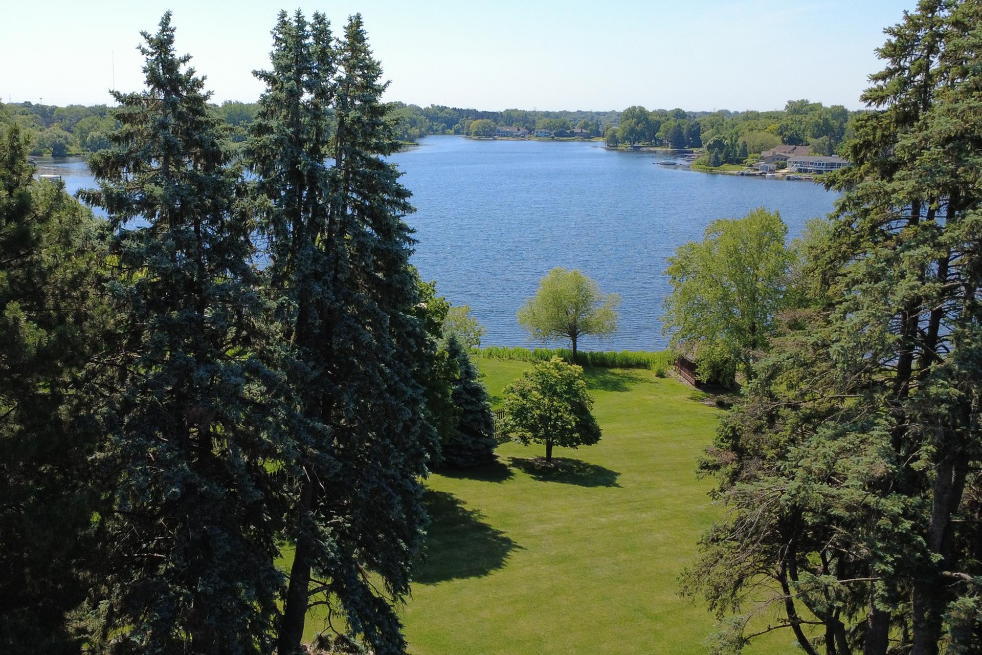 Waterfront lawn framed by tall evergreens, overlooking a lake and homes along the opposite shore.