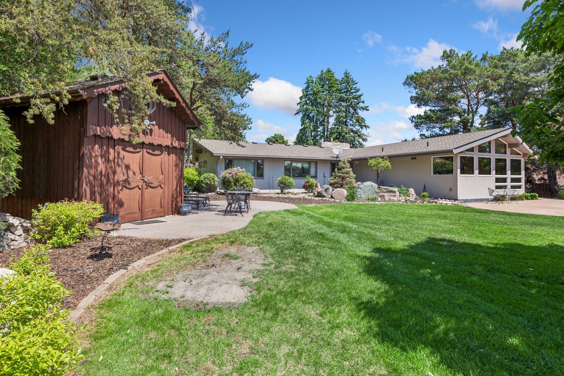 Single-story home with a wall of gabled windows, landscaped lawn, patio, and wooden outbuilding.