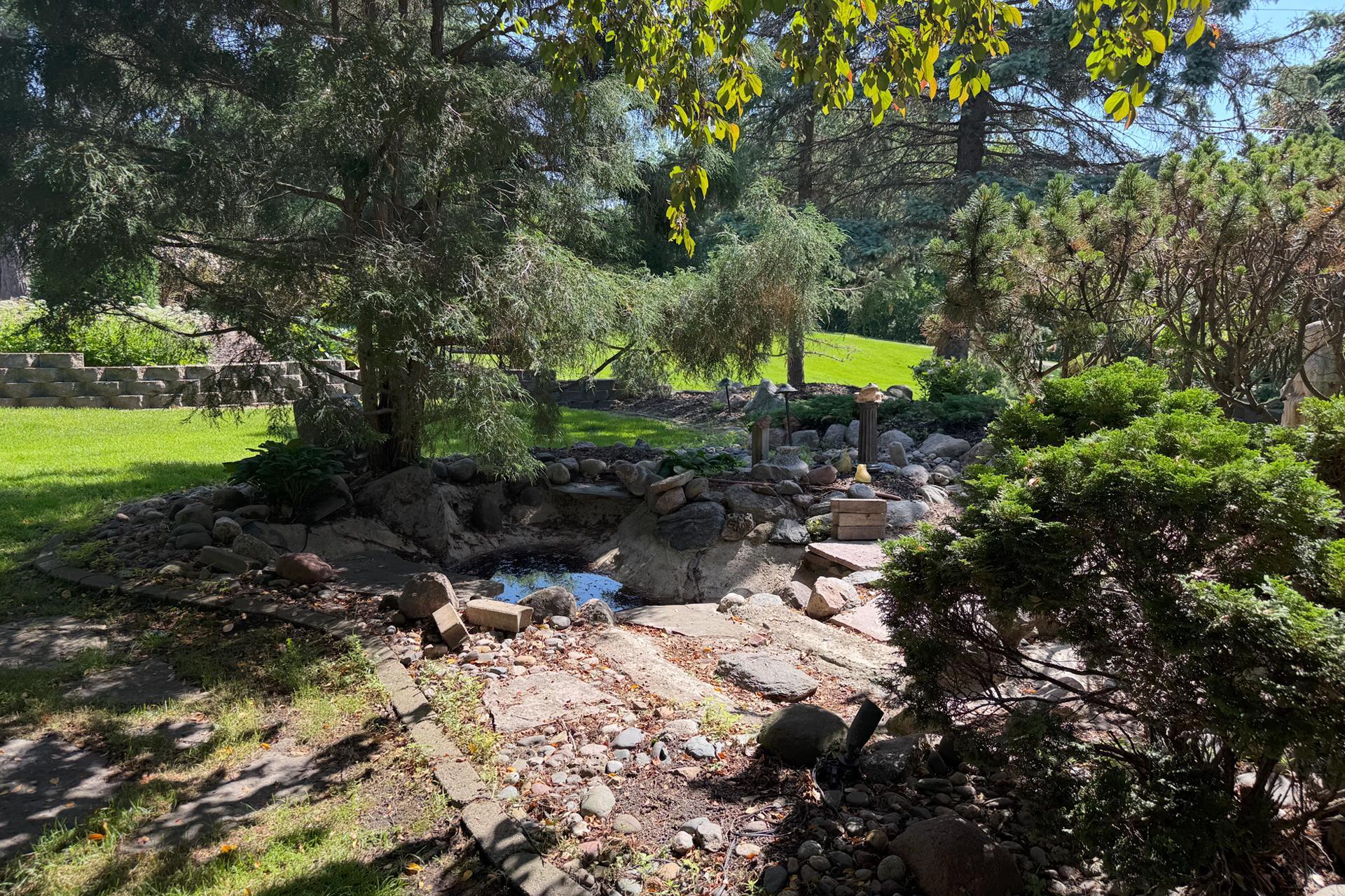 Landscaped backyard with a rock-edged pond, stone paths, mature evergreens, and a sunny lawn.