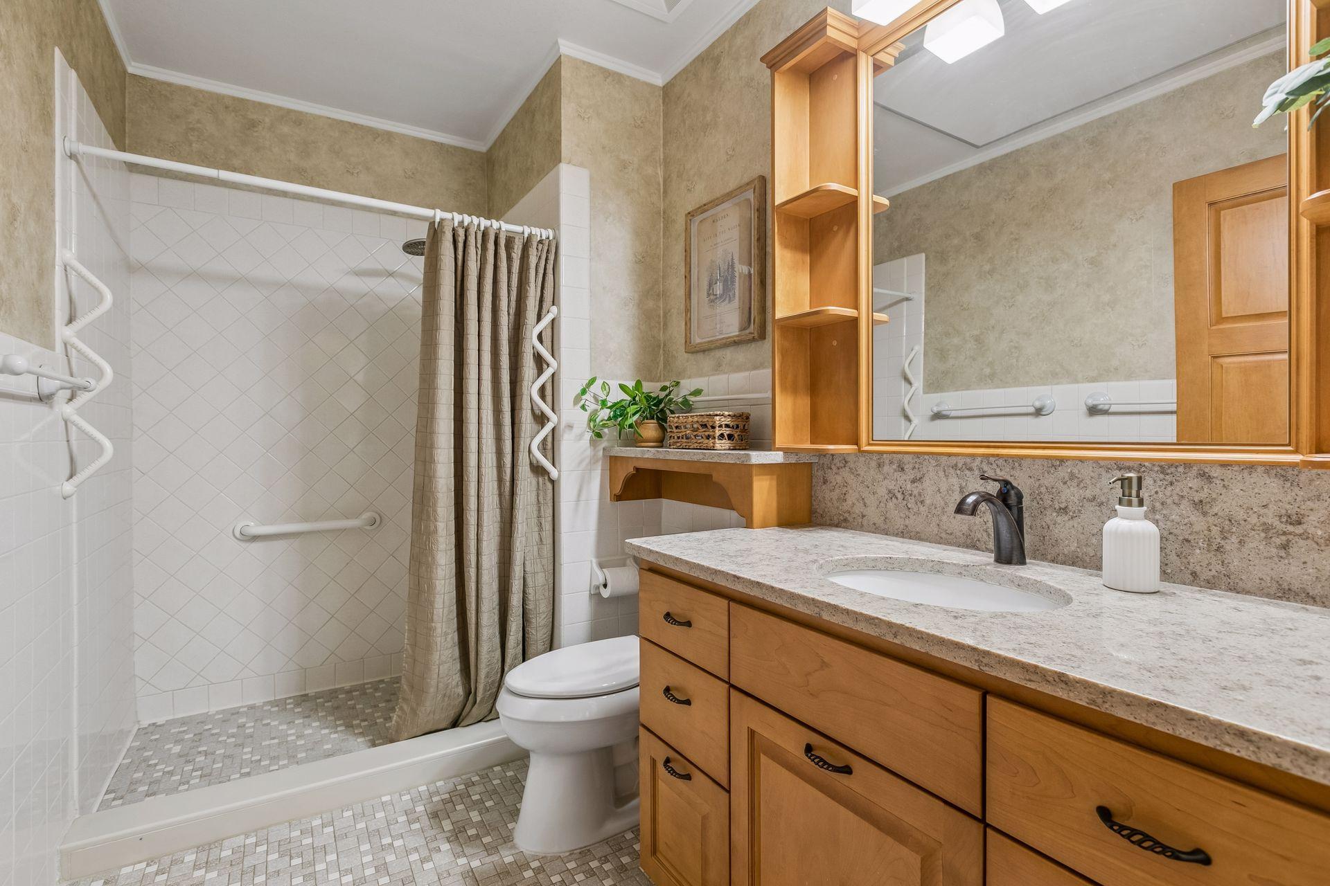 Bathroom with tiled shower, grab bars, toilet, and wood vanity beneath a large mirror.