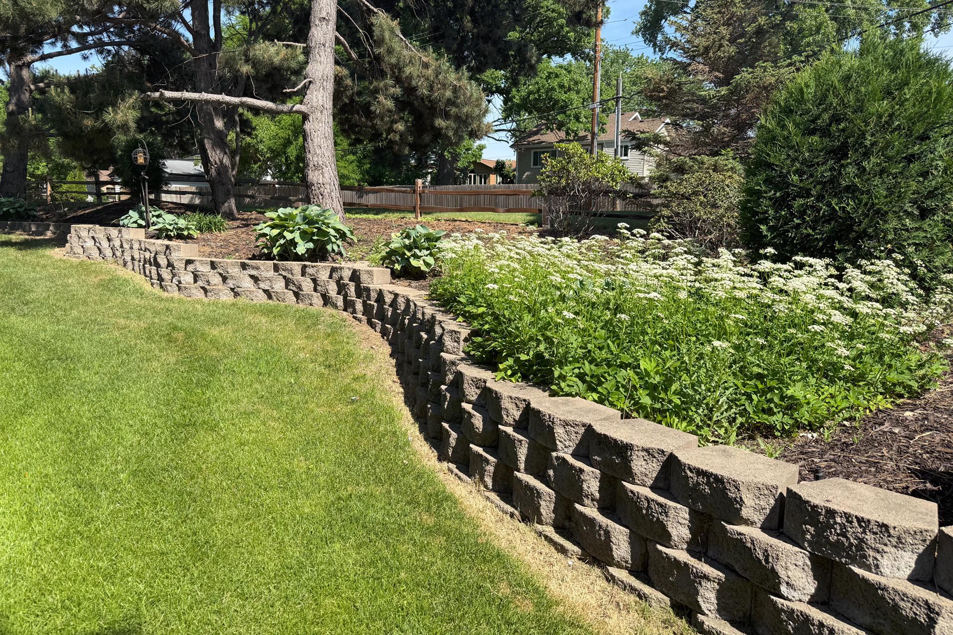 Landscaped yard with a curved stone retaining wall, flowering garden beds, and mature trees.
