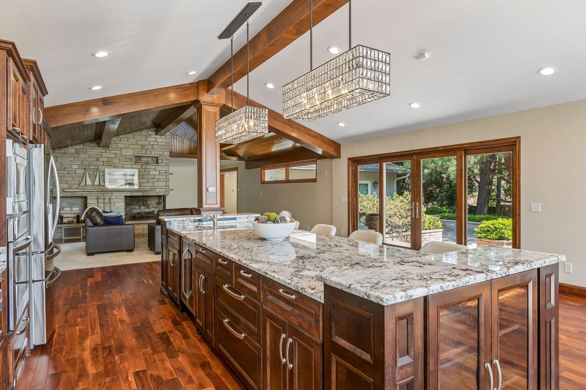 Open-plan kitchen with granite island, wood beams, stone fireplace, and glass doors to the patio.