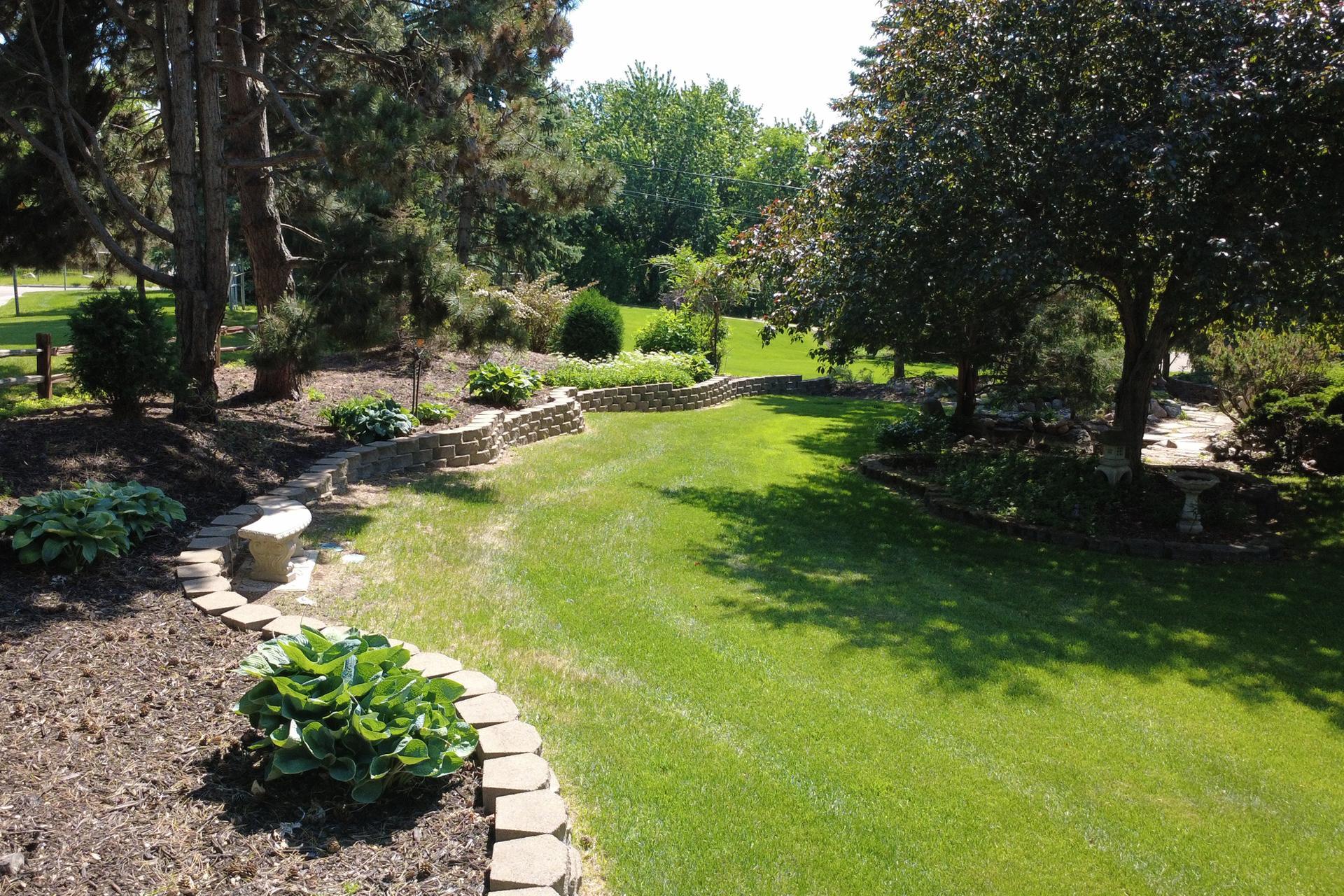Landscaped yard with a broad lawn, curved stone retaining walls, mature trees, and garden beds.