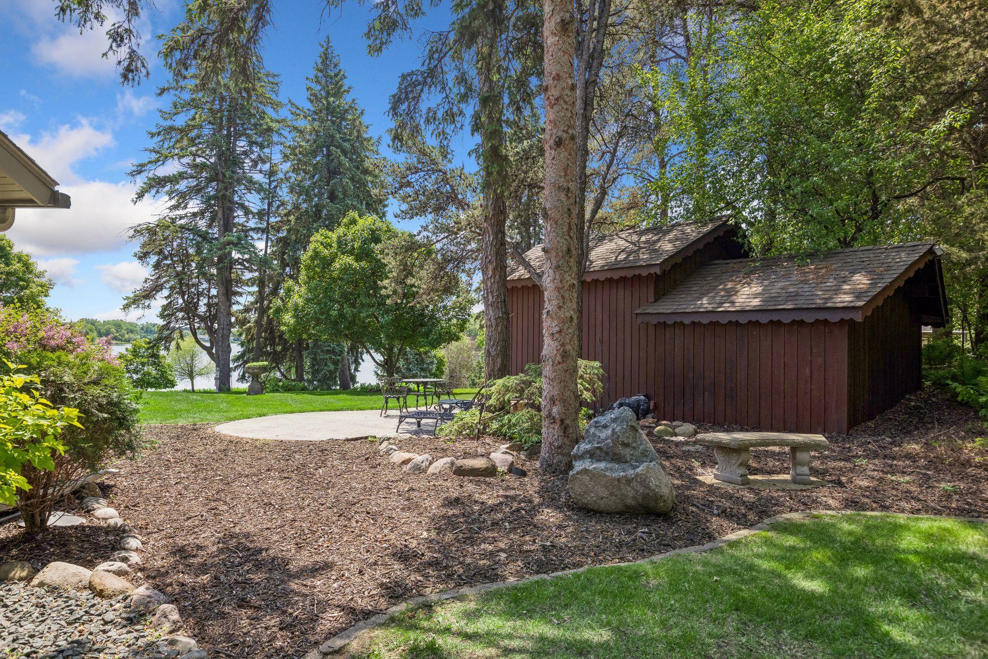 Lakeside backyard with a wooden shed, patio seating, mature trees, and a lake view.