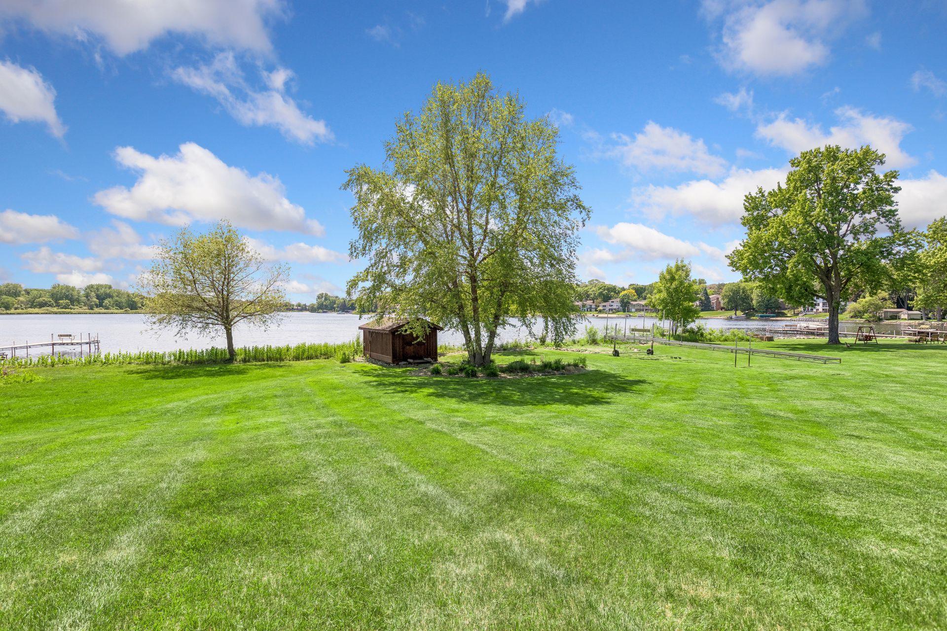 Expansive lakeside lawn with mature trees, a small shed, and docks along the water.