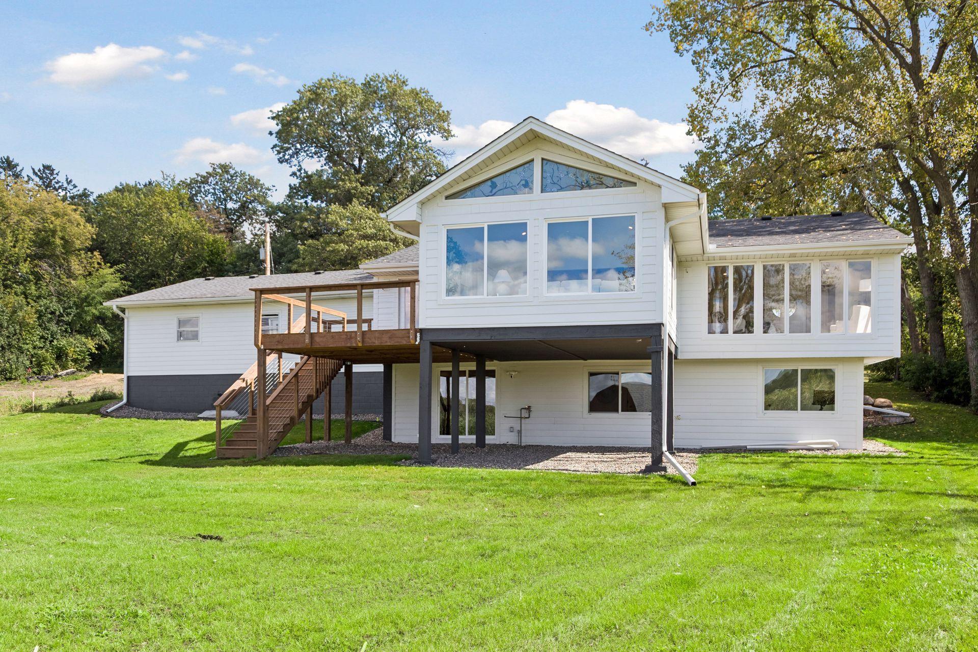 White split-level home with a raised wooden deck and stairs, large windows, and a spacious lawn.