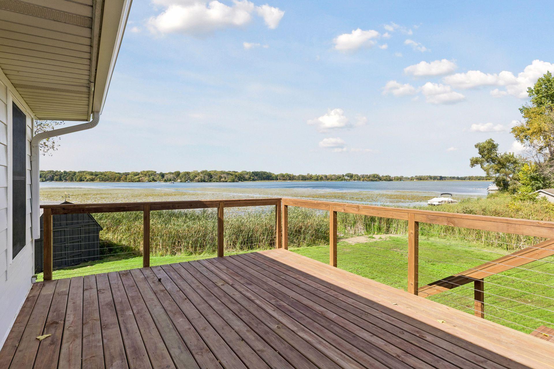 Wooden deck overlooking a lake, with cable railing, shoreline reeds, and a small boat.