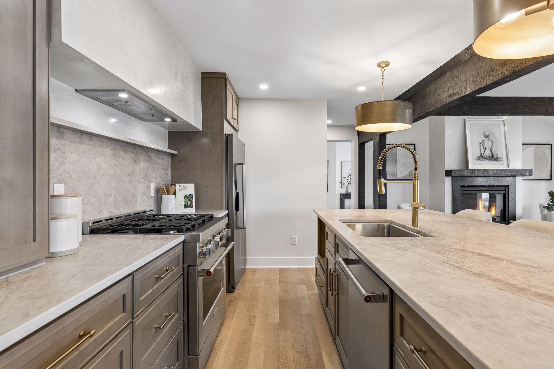 Renovated kitchen with a stone island, brass faucet, gas range, and exposed beams beside a fireplace.