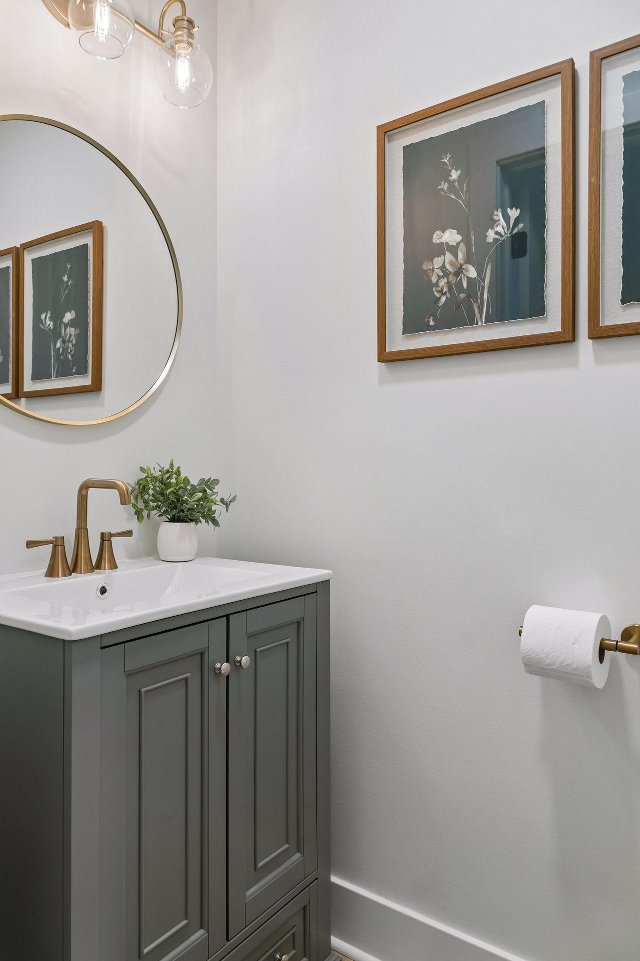 Bathroom vanity with a gray-green cabinet, brass faucet, round mirror, and framed botanical prints.