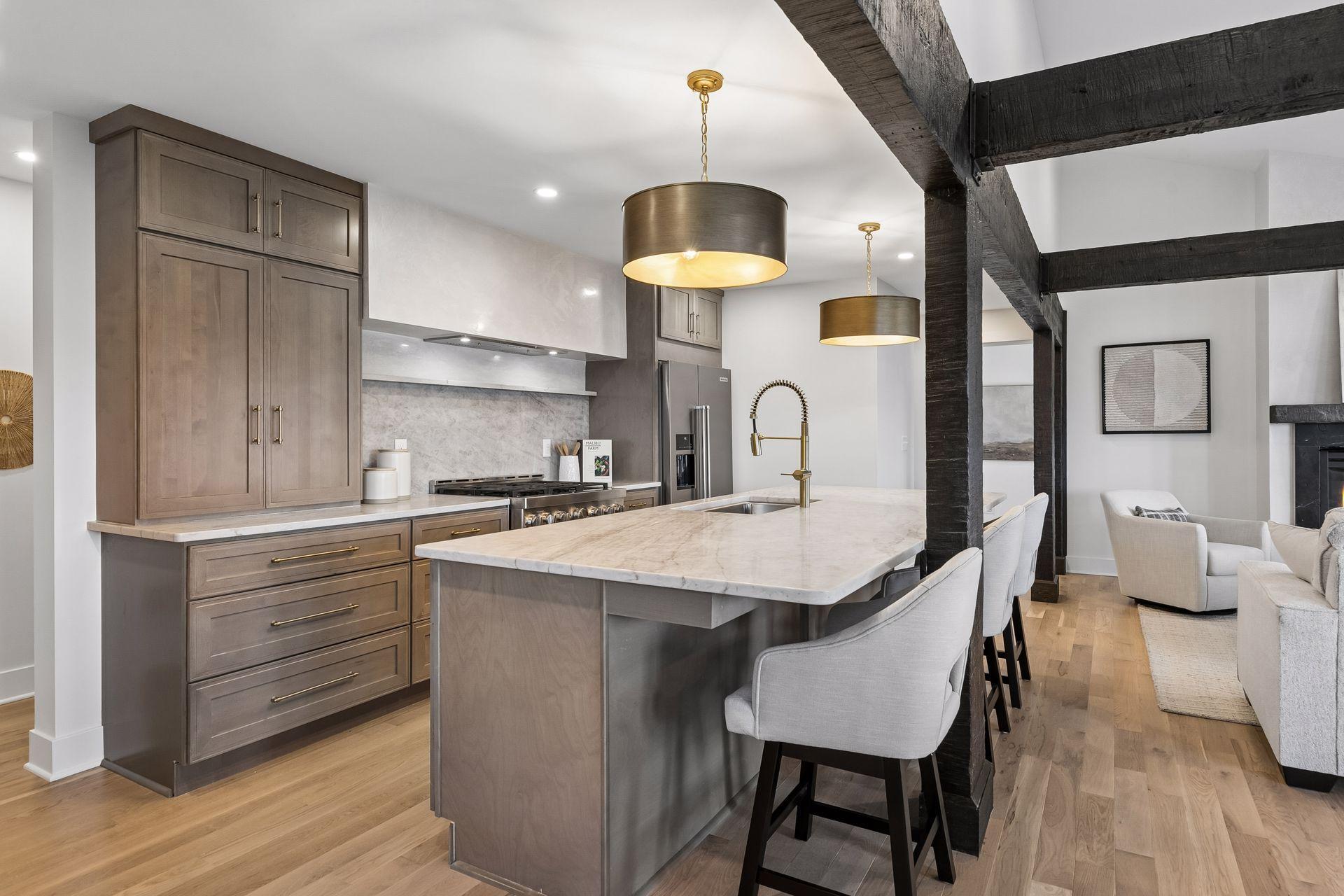 Kitchen with a large stone island, gray-brown cabinets, brass fixtures, and dark beams overlooking the living area.