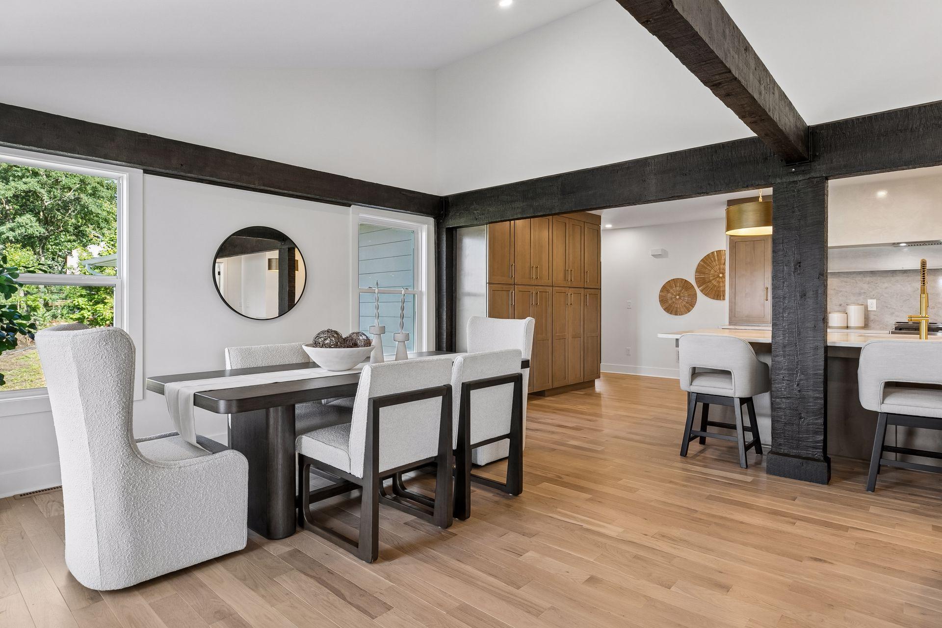 Dining area with dark exposed beams, light upholstered chairs, and an open view into the kitchen.