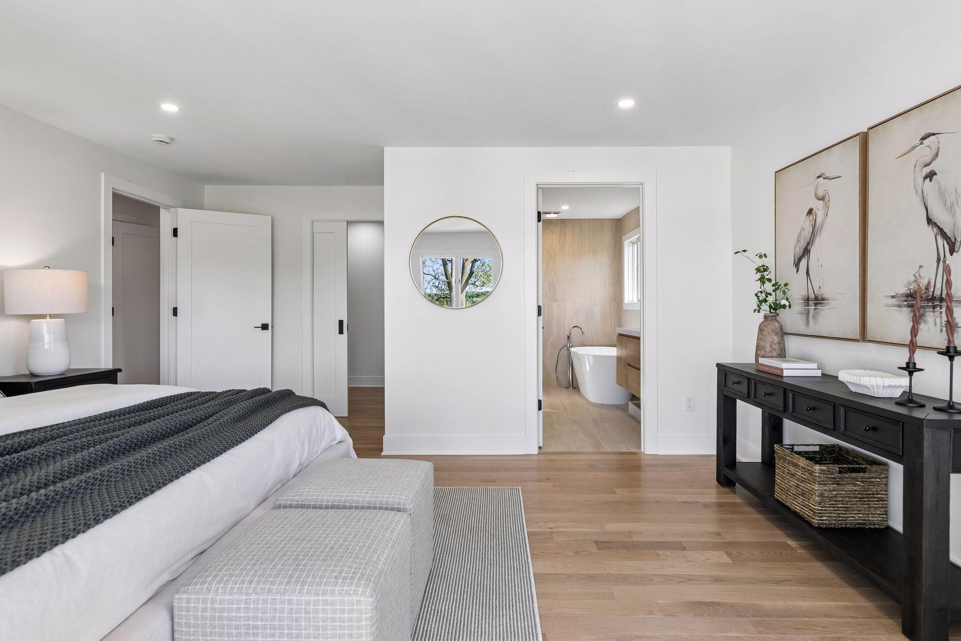 Staged bedroom with a view into an ensuite bathroom and light wood flooring.