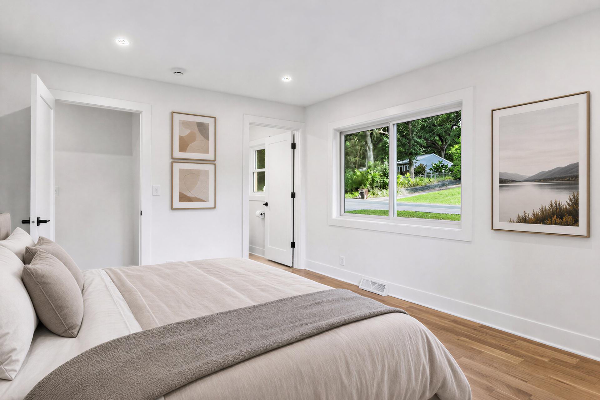 Bright staged bedroom with a large bed, hardwood floors, and a wide window overlooking greenery.