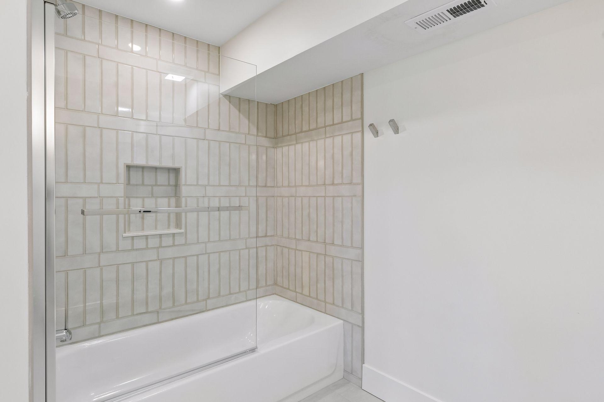 Bathroom tub and shower with glass screen and pale tiled walls.