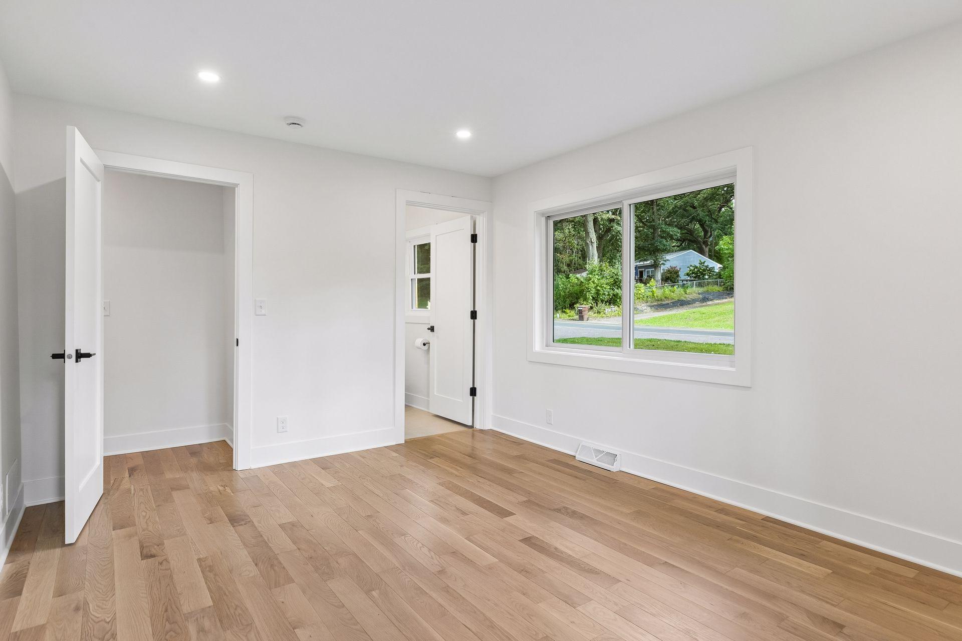 Empty bedroom with light wood floors, a closet, and a large window overlooking greenery.