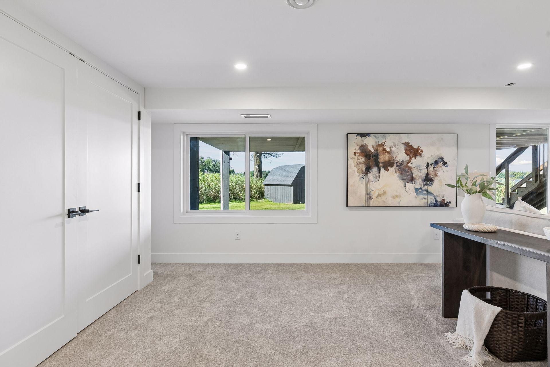 Carpeted room with a wide window overlooking a lawn and outbuilding, abstract wall art, and a dark console table.