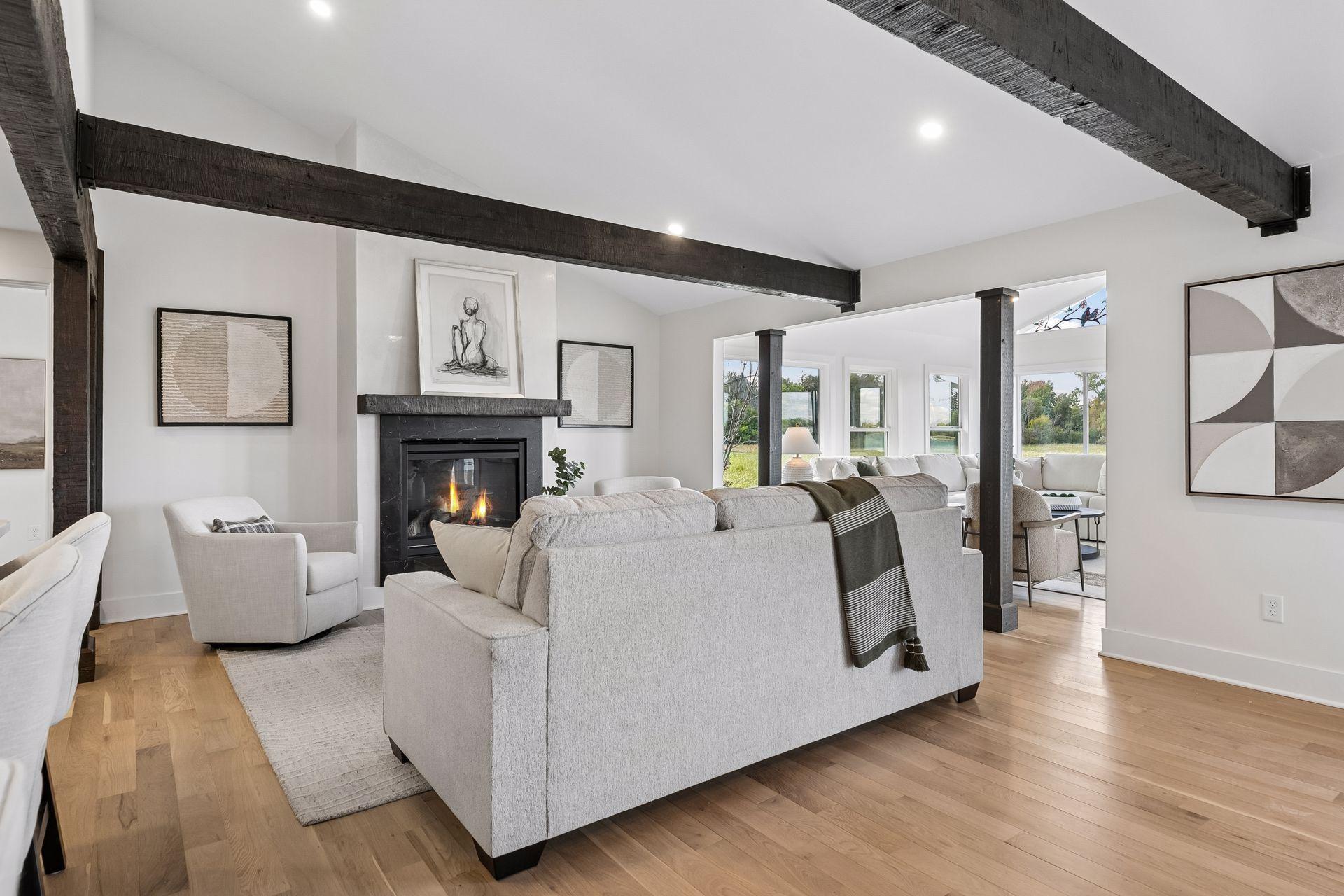 Living room with a black fireplace, exposed dark beams, and pale seating beside large windows.