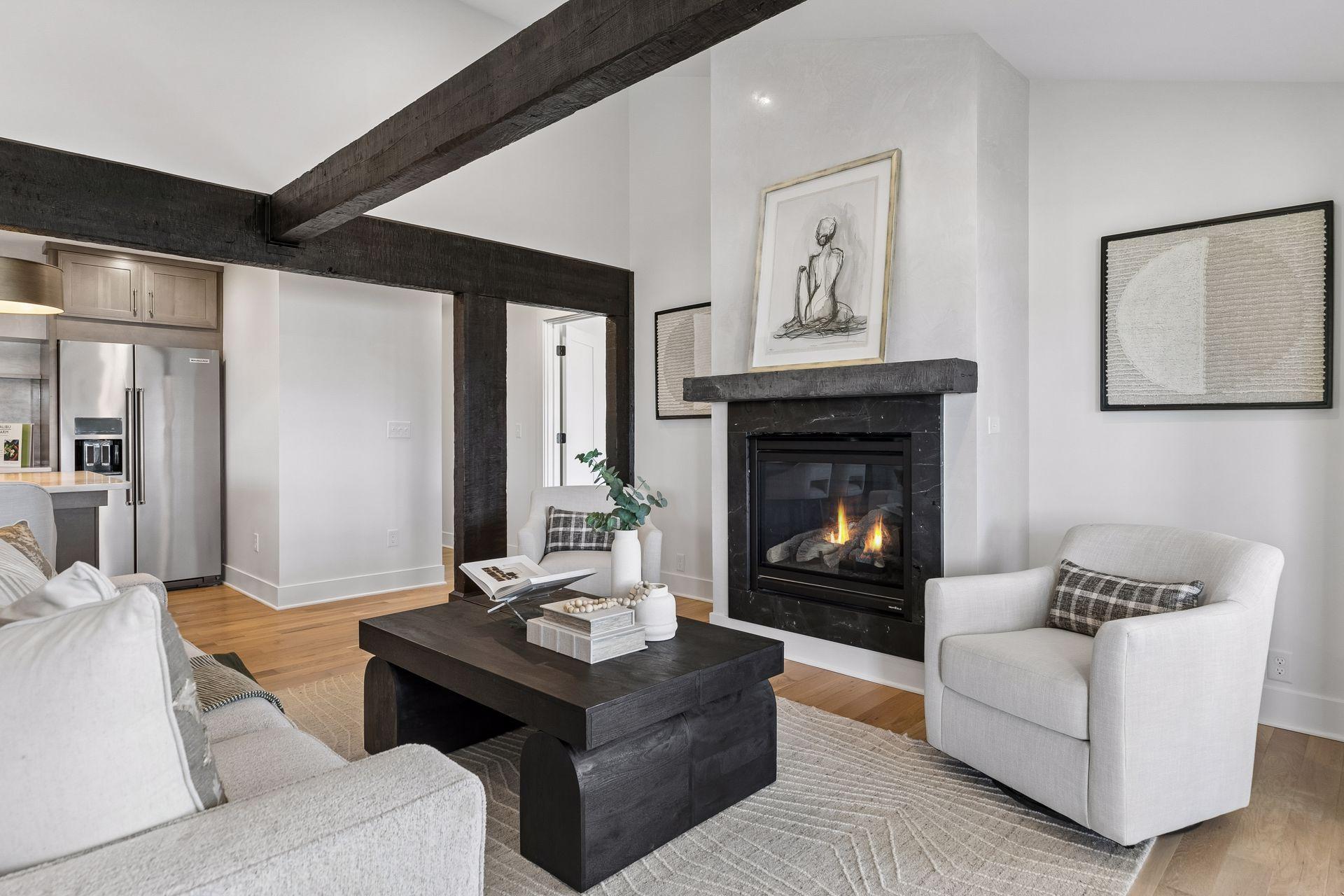 Living room with a lit fireplace, exposed dark beams, and white seating beside the kitchen.