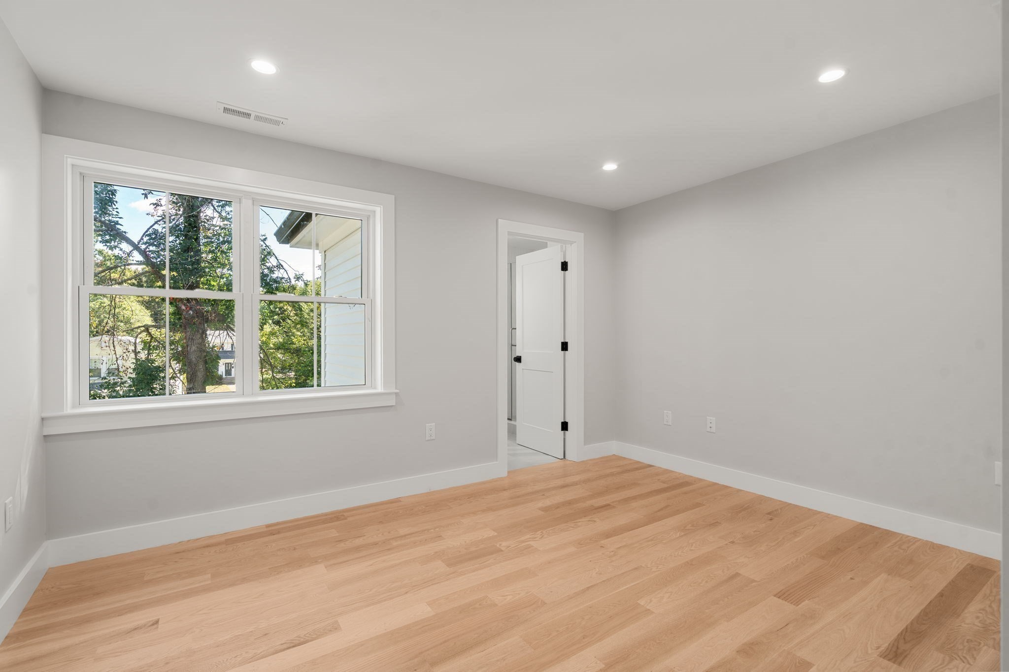 Empty room with hardwood floors, recessed lights, and a large window overlooking trees.