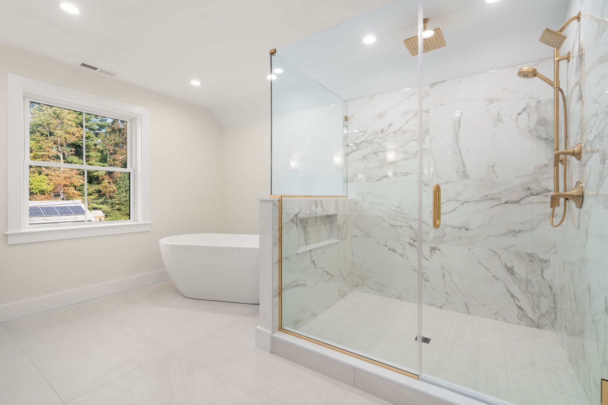Bathroom with a freestanding soaking tub and glass shower with marble-look walls and gold fixtures.