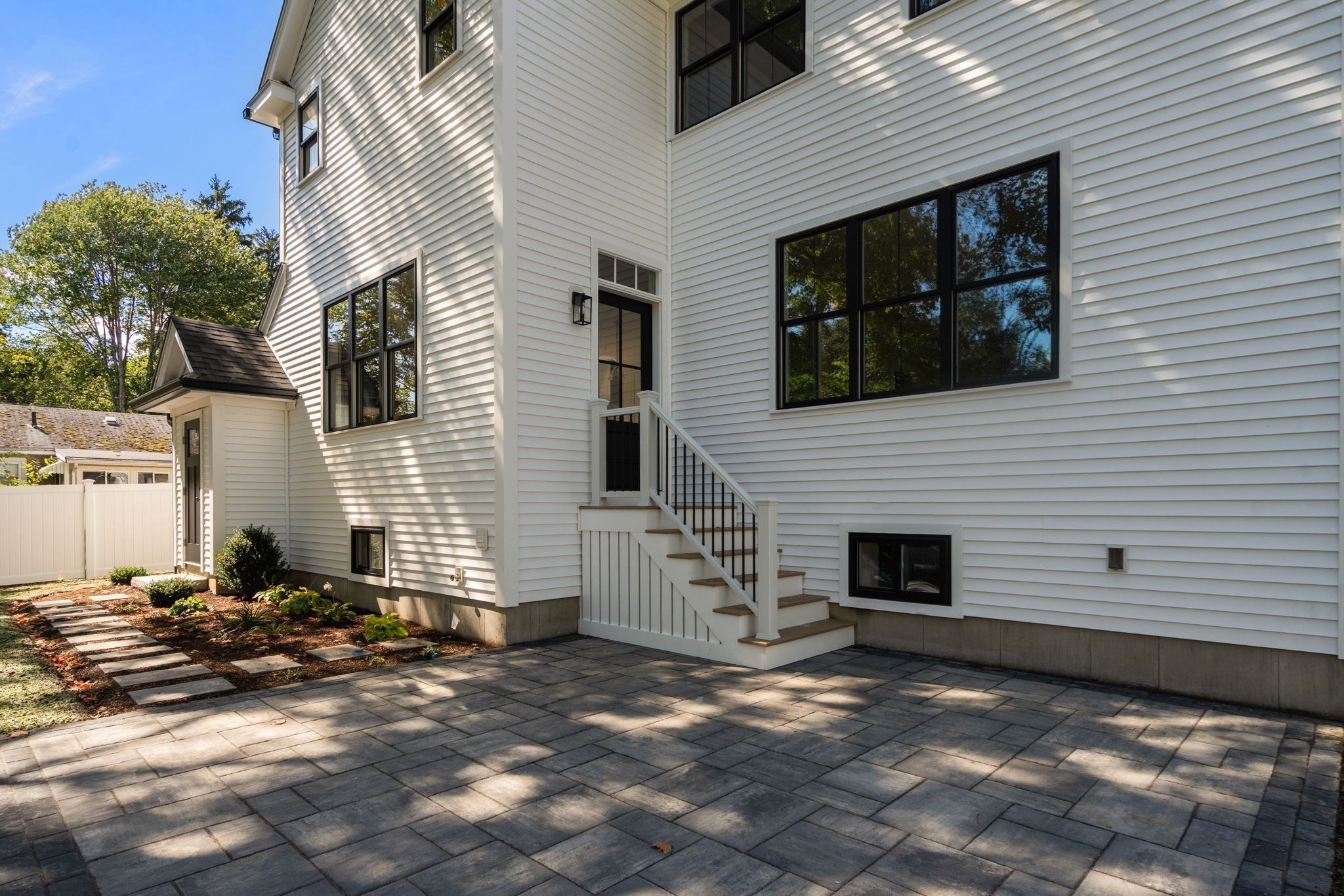 White-sided home with black-framed windows and steps opening onto a broad stone-paver patio.