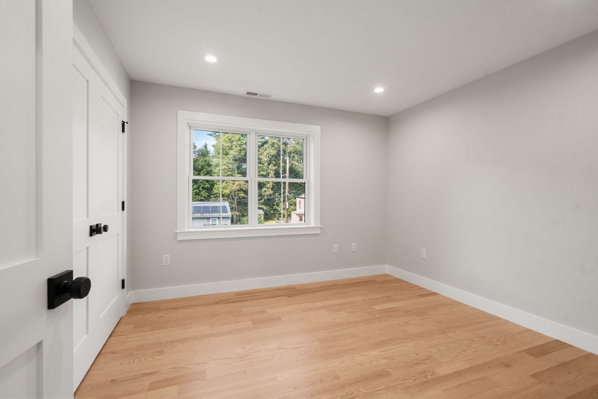 Empty room with light hardwood floors, a white paneled door, and a window overlooking trees.