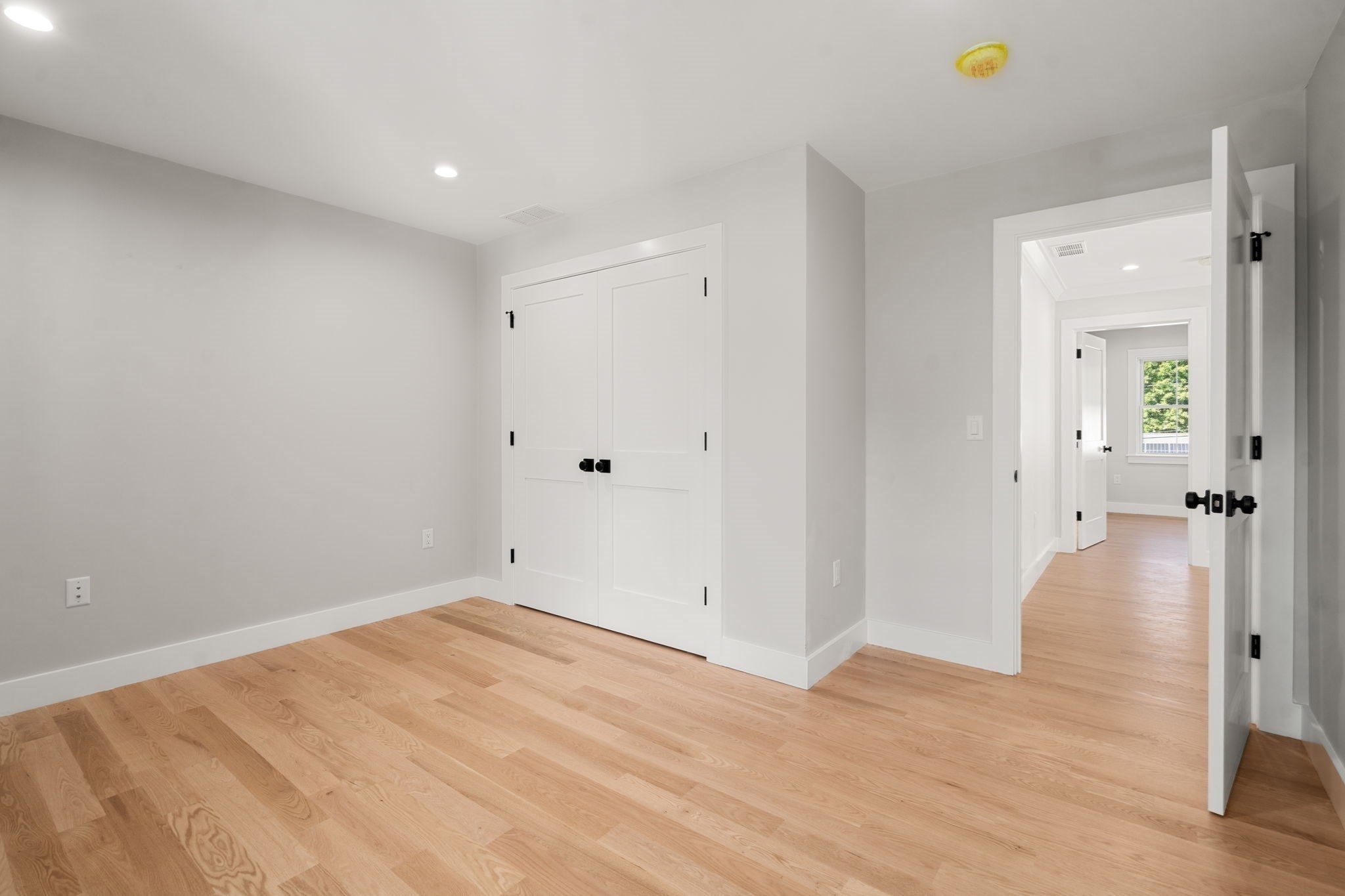 Empty room with light oak floors, white closet doors, and an open doorway to a room with a window.