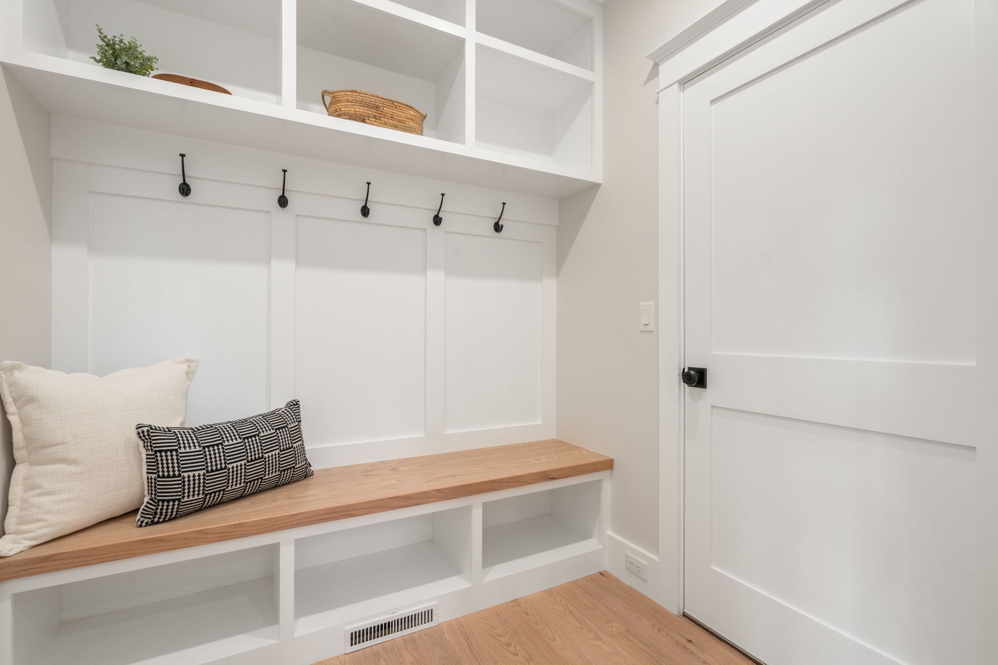 Mudroom with a built-in wooden bench, black hooks, open cubbies, and a white paneled door.