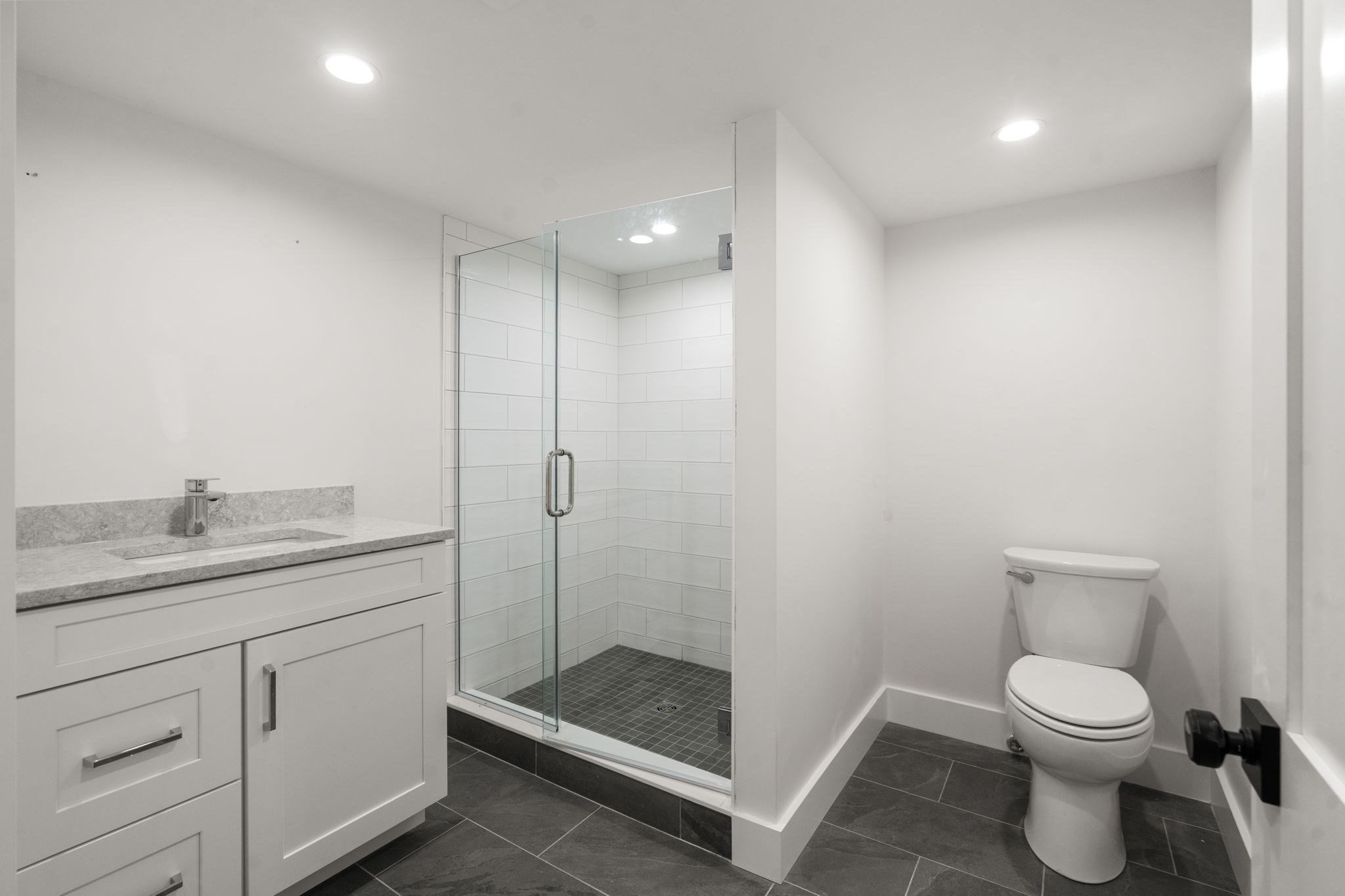 Modern bathroom with a glass walk-in shower, gray vanity, and dark tile floor.