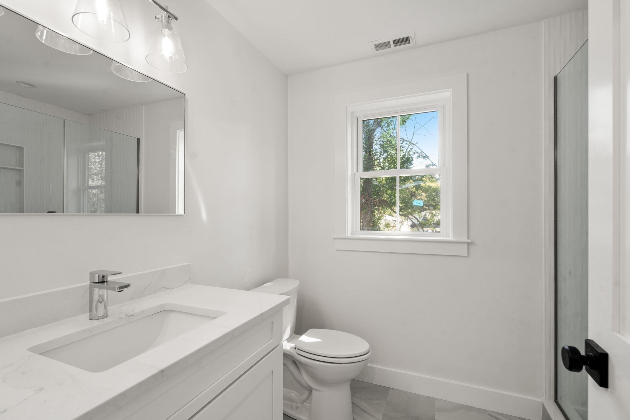 Bright white bathroom with a marble-look vanity, window, toilet, and glass shower enclosure.