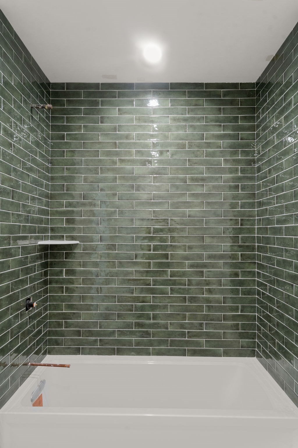 Bathtub beneath dark green subway-tiled shower walls with exposed plumbing fittings.