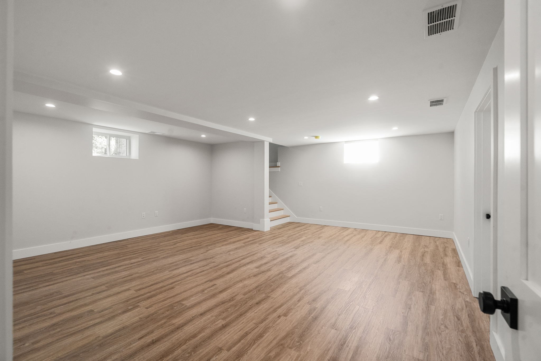 Vacant finished basement with wood-look floors, recessed lights, high windows, and stairs.