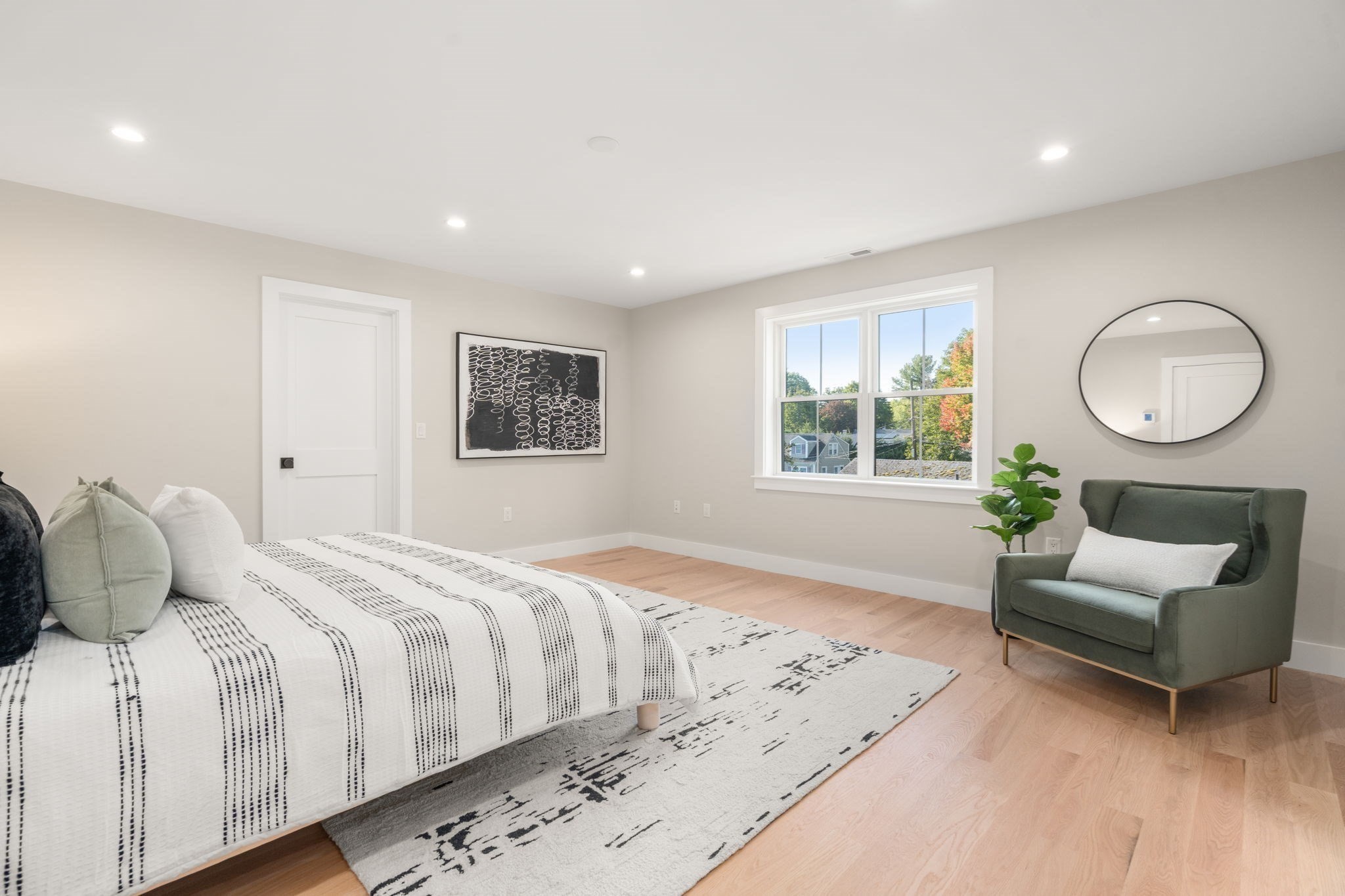 Staged bedroom with patterned white bedding, pale wood floors, and a green chair beside the window.
