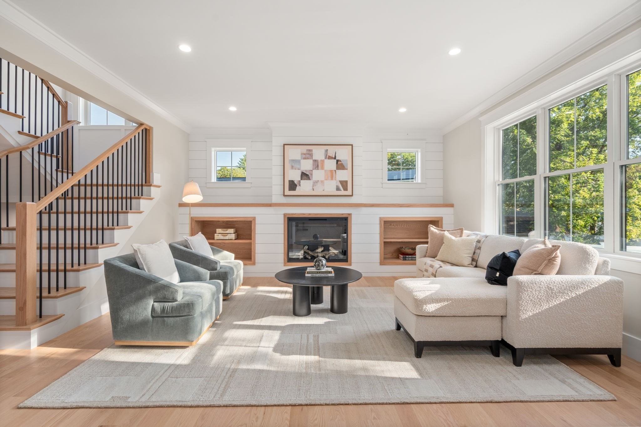 Staged living room with a shiplap fireplace, built-in shelves, staircase, and large sunny windows.