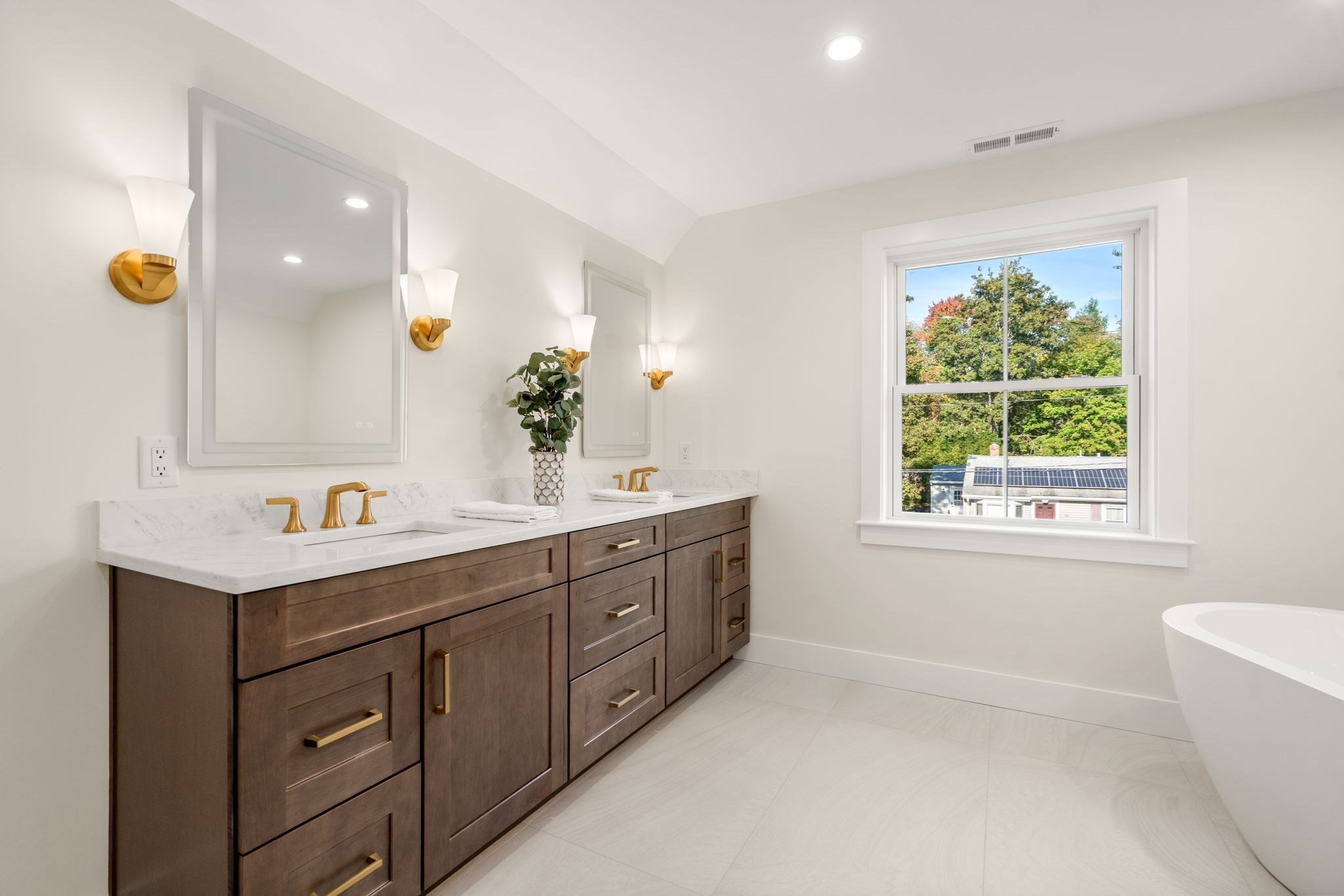 Bathroom with dark wood double vanity, marble counters, brass fixtures, and a window overlooking trees.