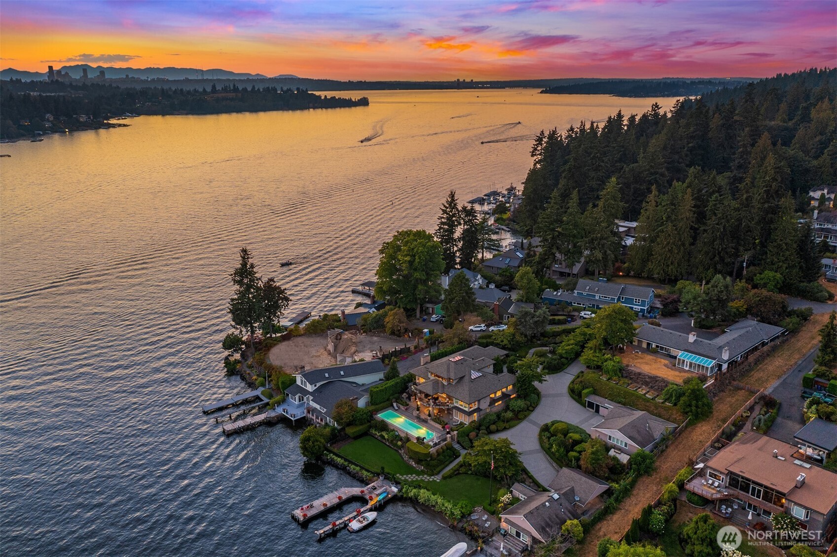 Waterfront homes and docks beside a wooded shoreline at sunset, with a pool and distant city skyline.