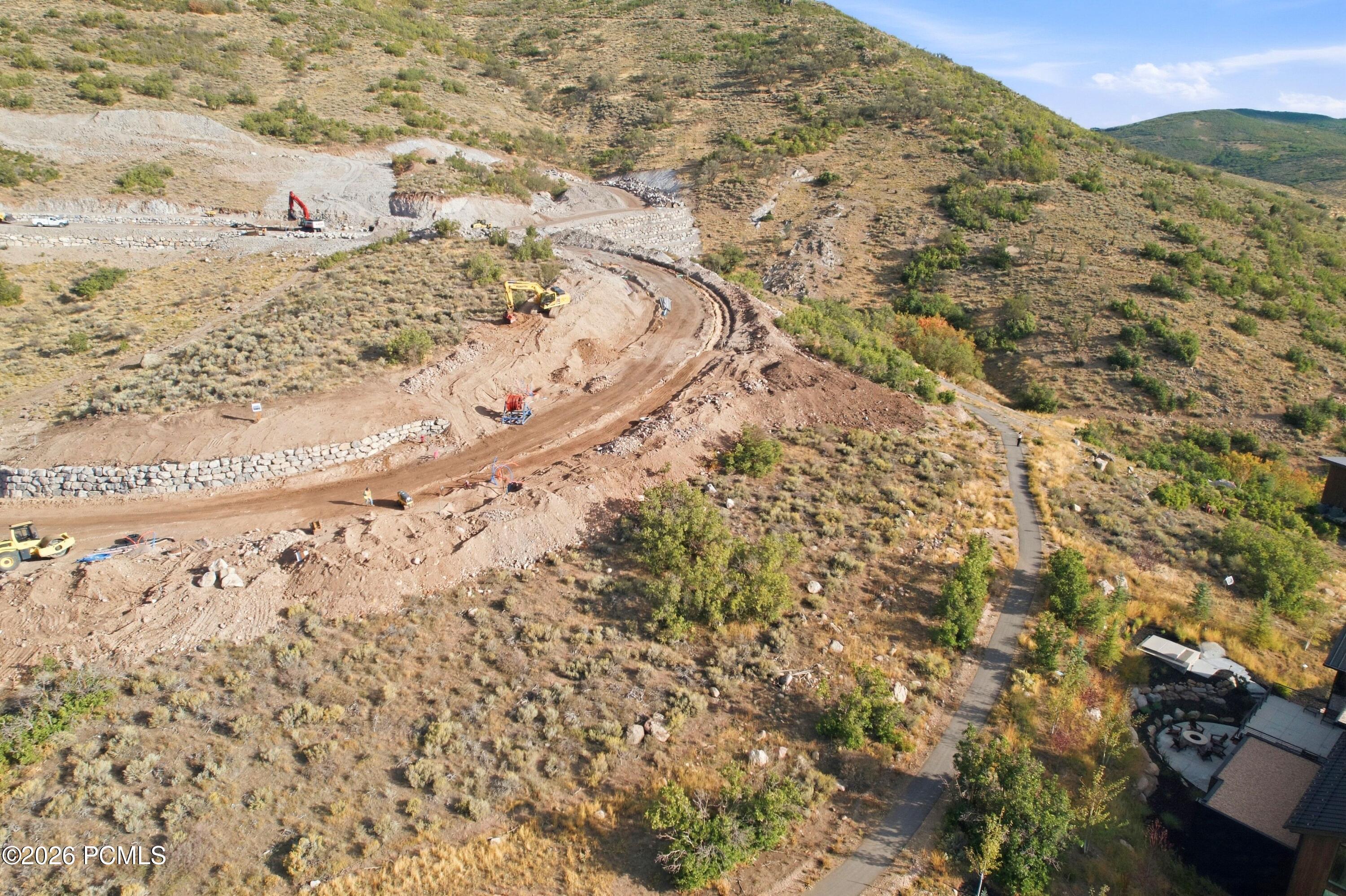 Hillside construction site with a winding graded road, excavators, and stone retaining walls.