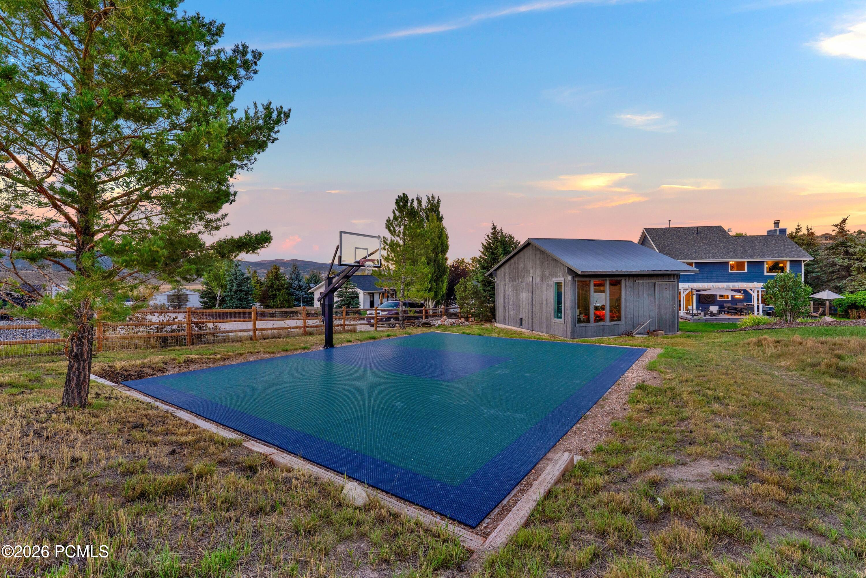 Backyard basketball court beside a gray outbuilding and blue house at sunset.