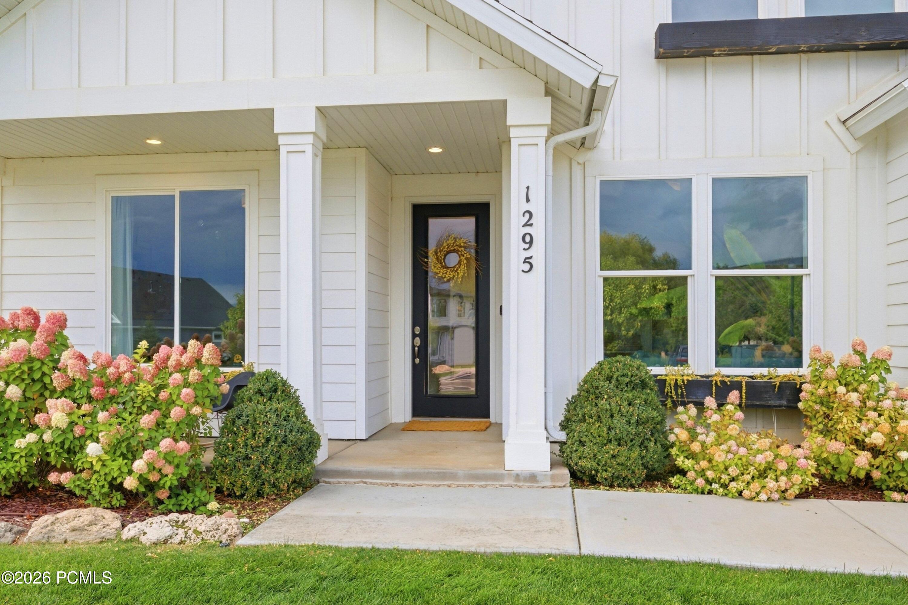 White home’s covered entry with a black glass door and pink flowering shrubs.
