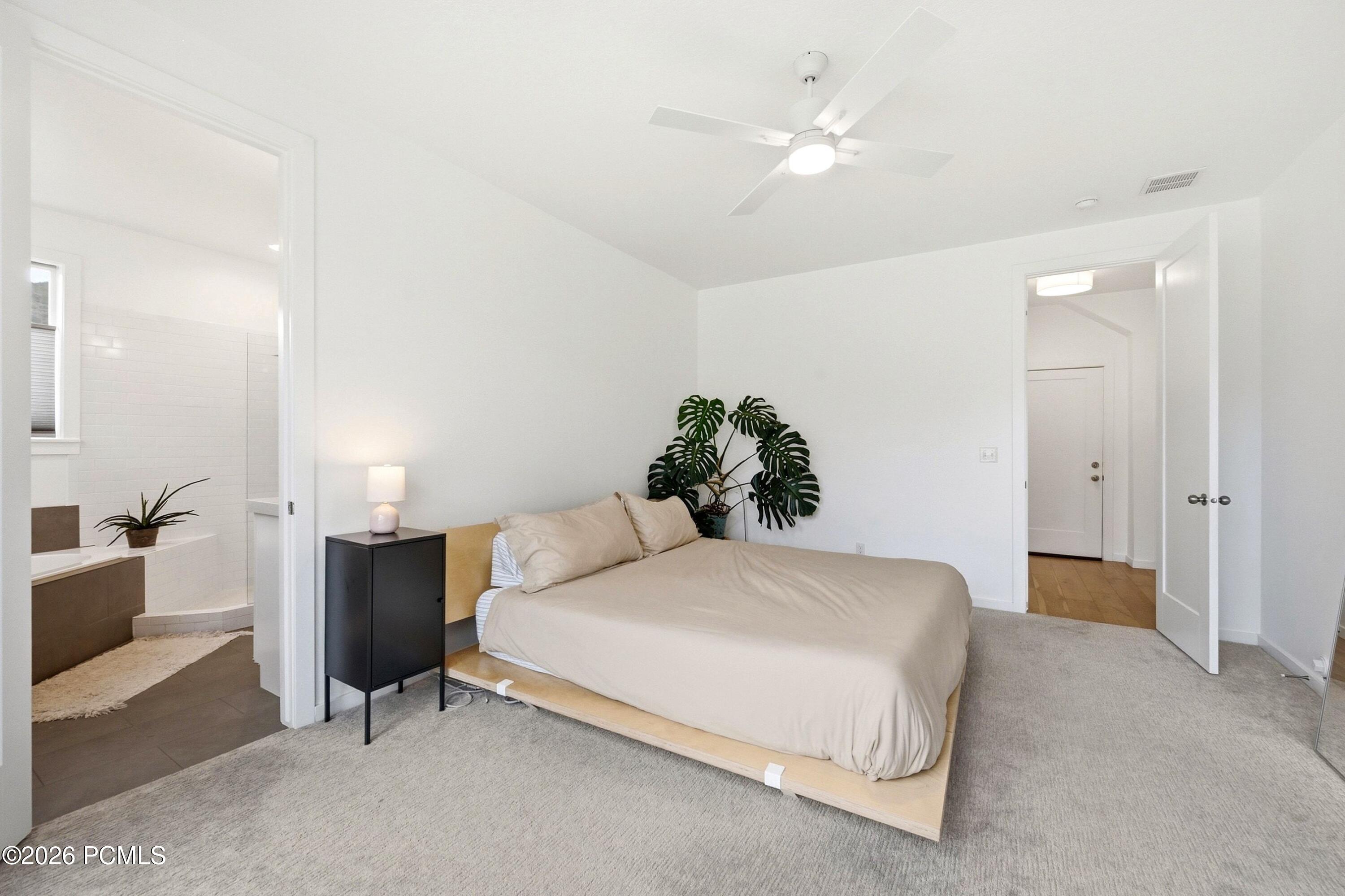 Staged bedroom with a low platform bed and ceiling fan beside an adjoining tiled bathroom.
