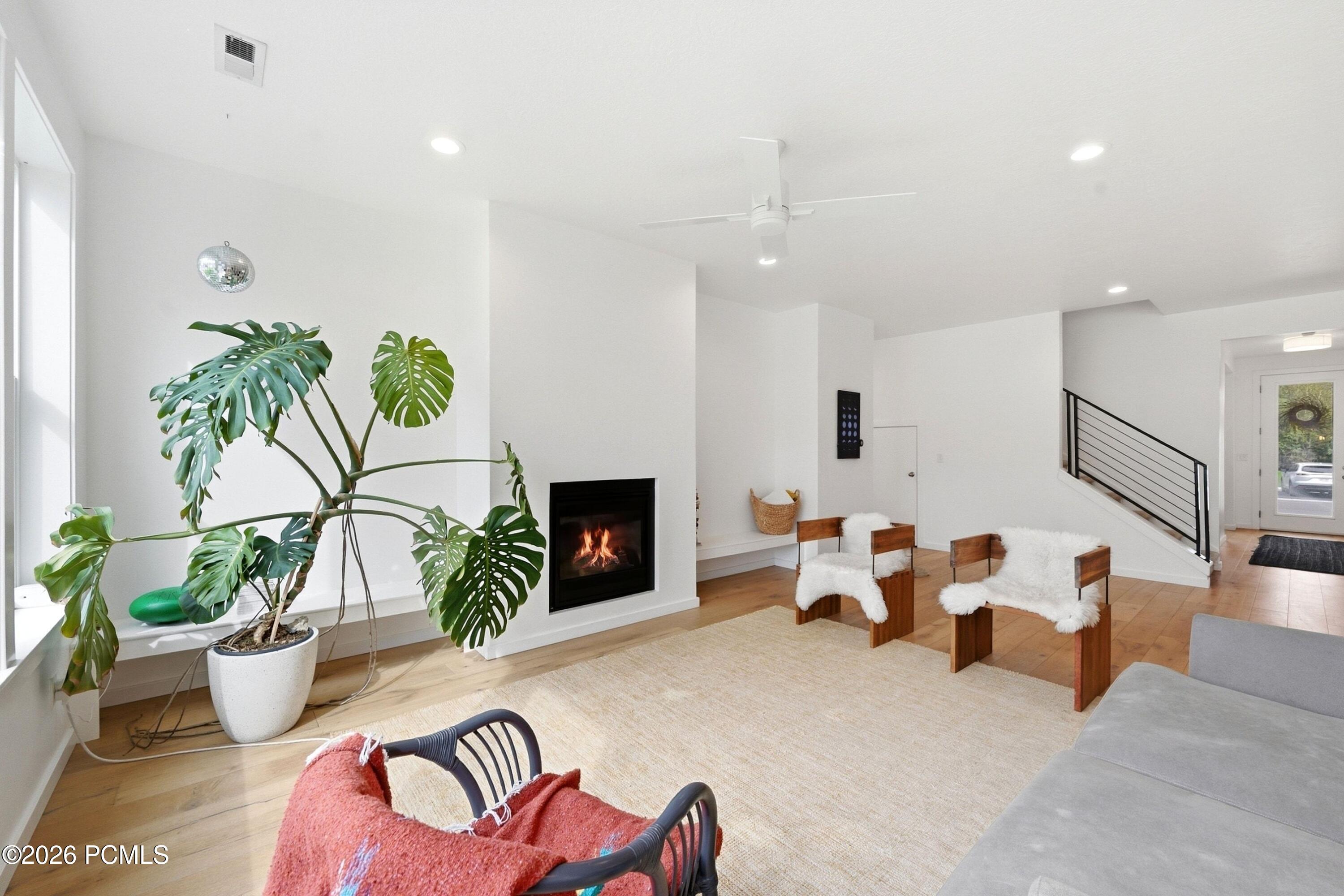Living room with a lit fireplace, large monstera plant, white-cushioned chairs, and a black-railed staircase.