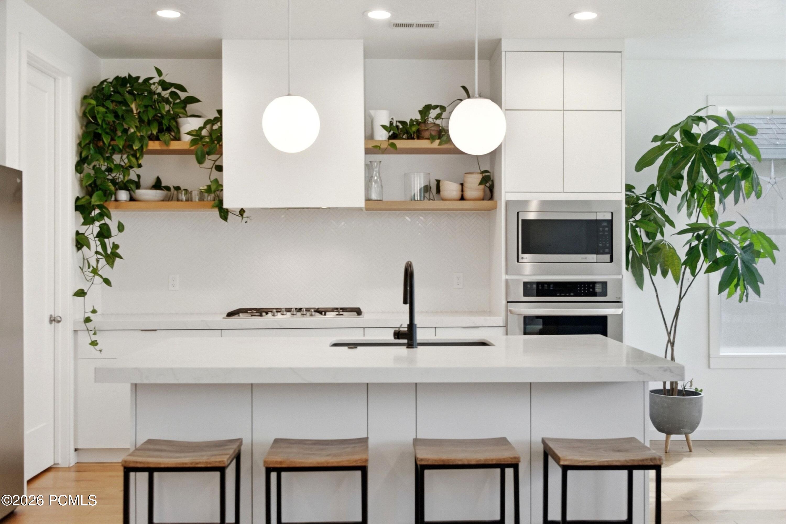 Modern white kitchen with a broad island, wooden stools, open plant-filled shelves, and built-in appliances.