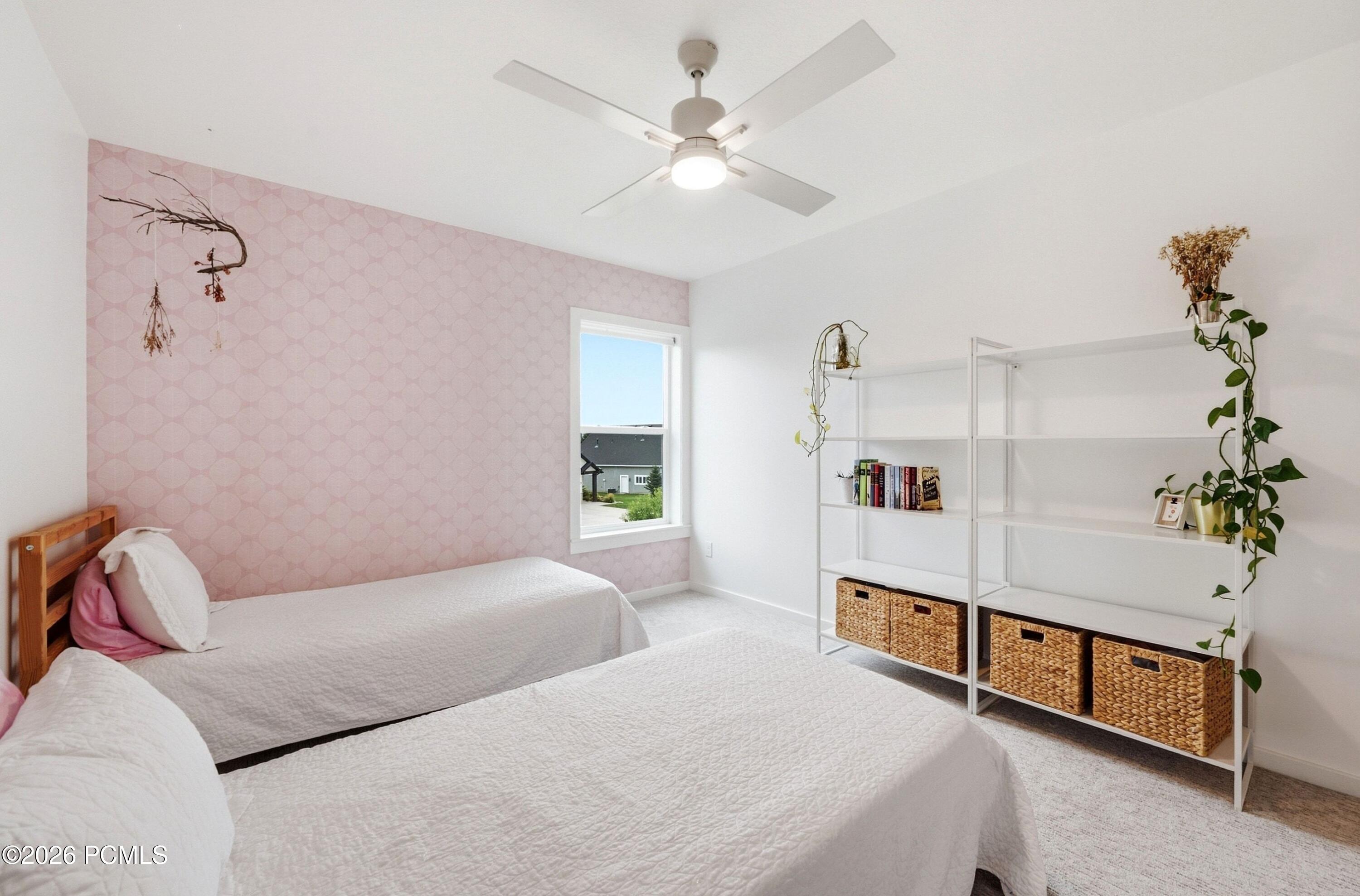 Bright bedroom with two white-covered beds, a pink patterned accent wall, and open shelving.