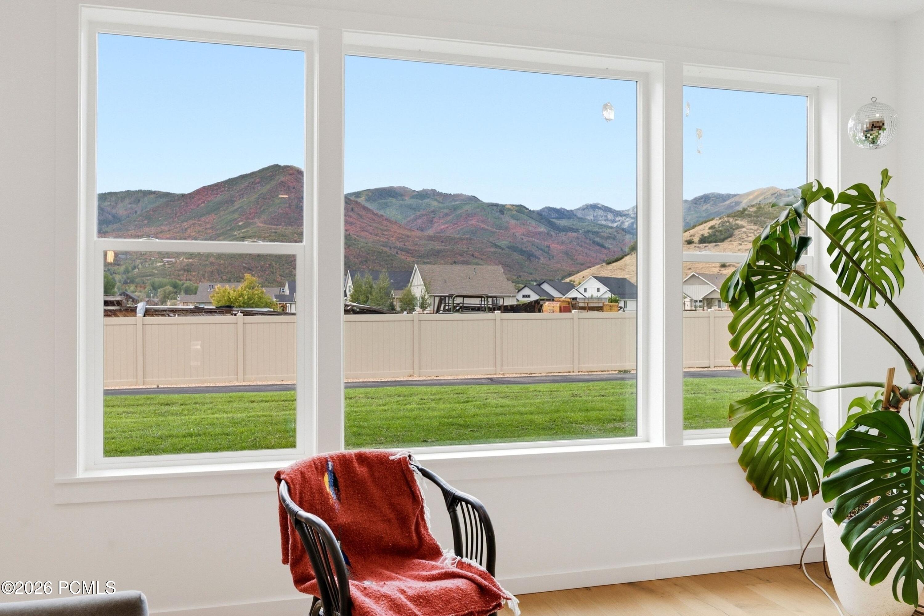 Large living room windows frame autumn-colored mountains beyond the lawn, with a chair and monstera plant nearby.