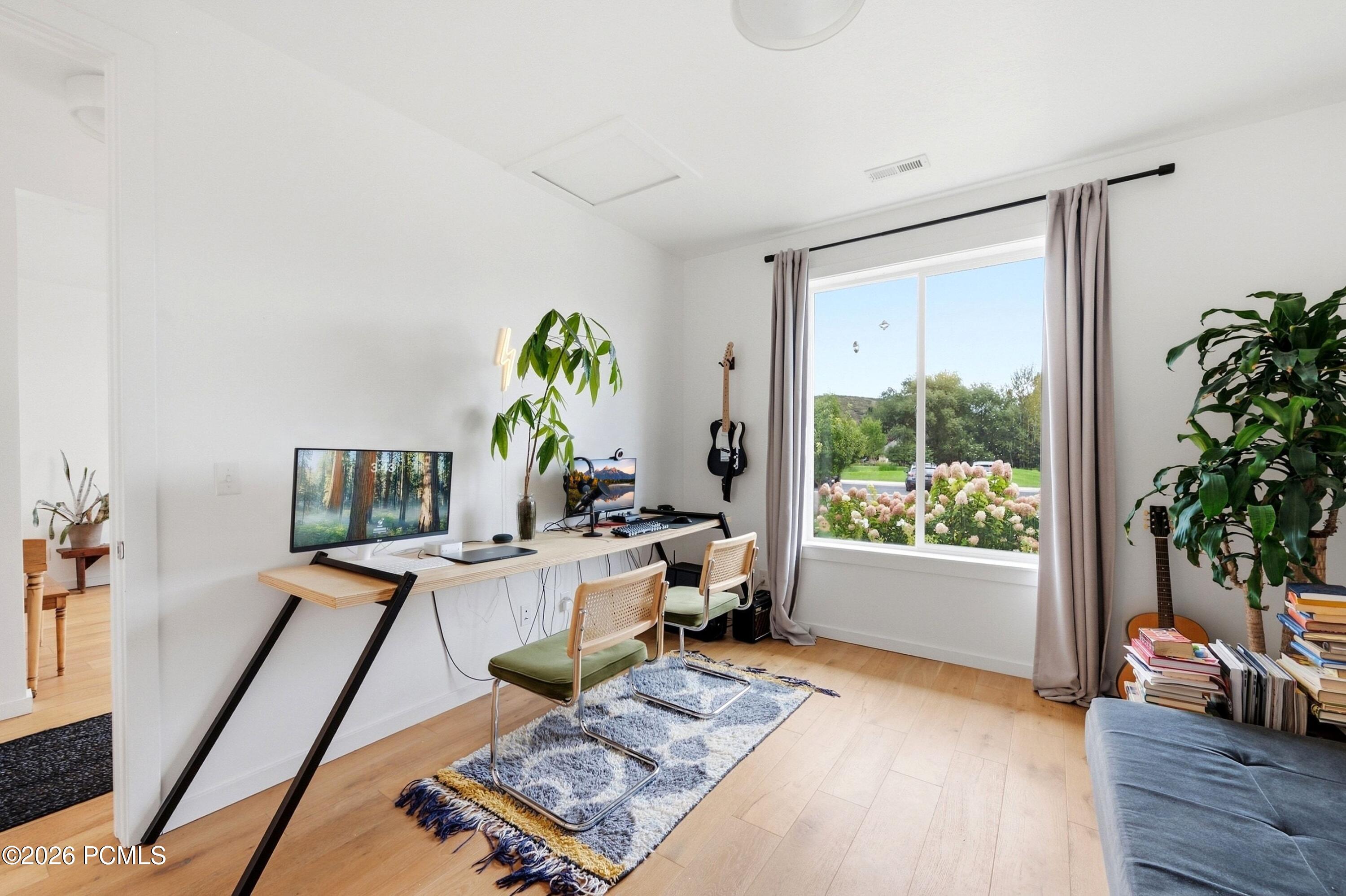 Furnished home office with computer desks, guitars, leafy plants, and a large window overlooking flowering shrubs.