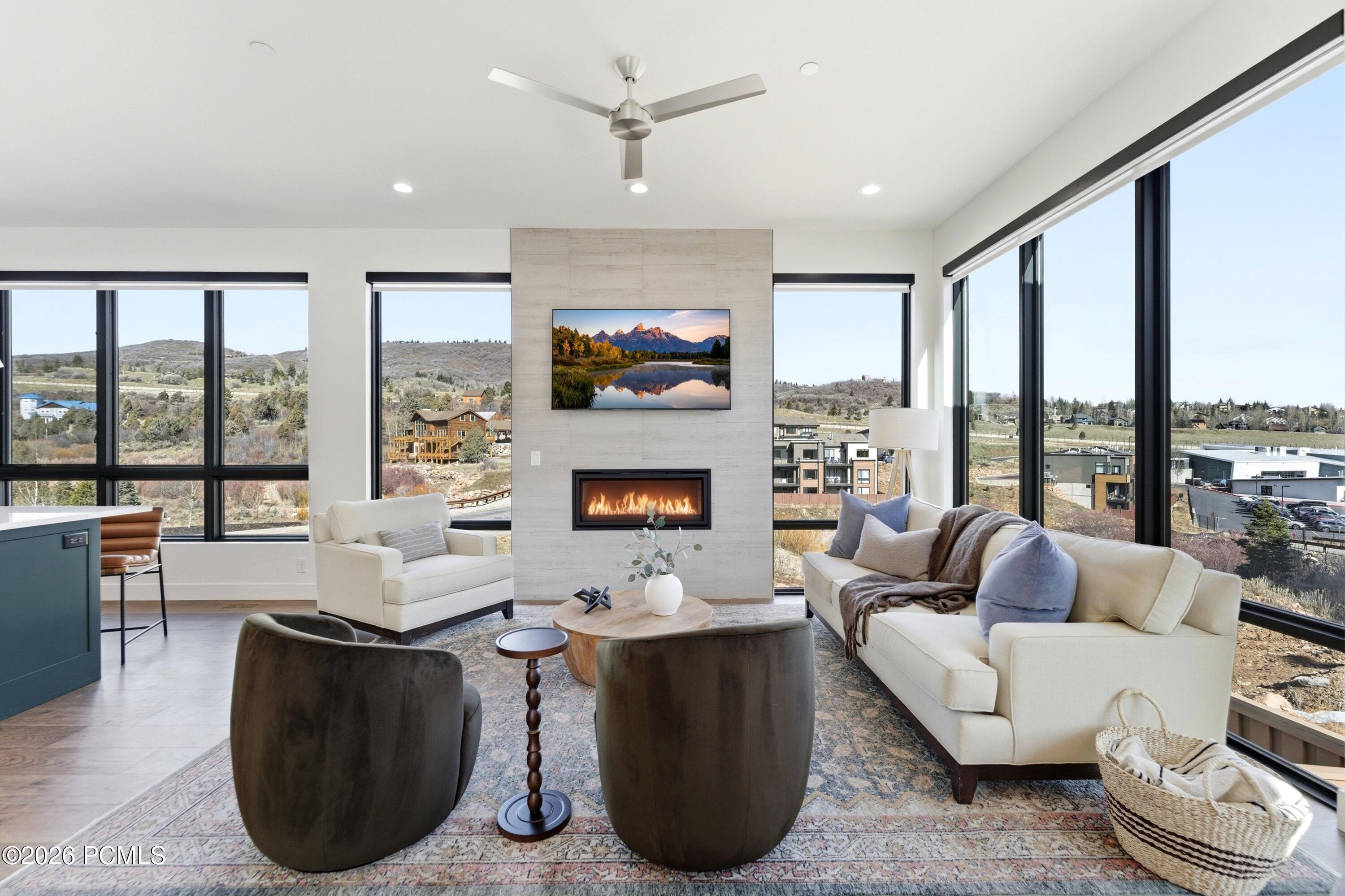 Furnished living room with a stone fireplace and expansive windows overlooking nearby homes and hills.