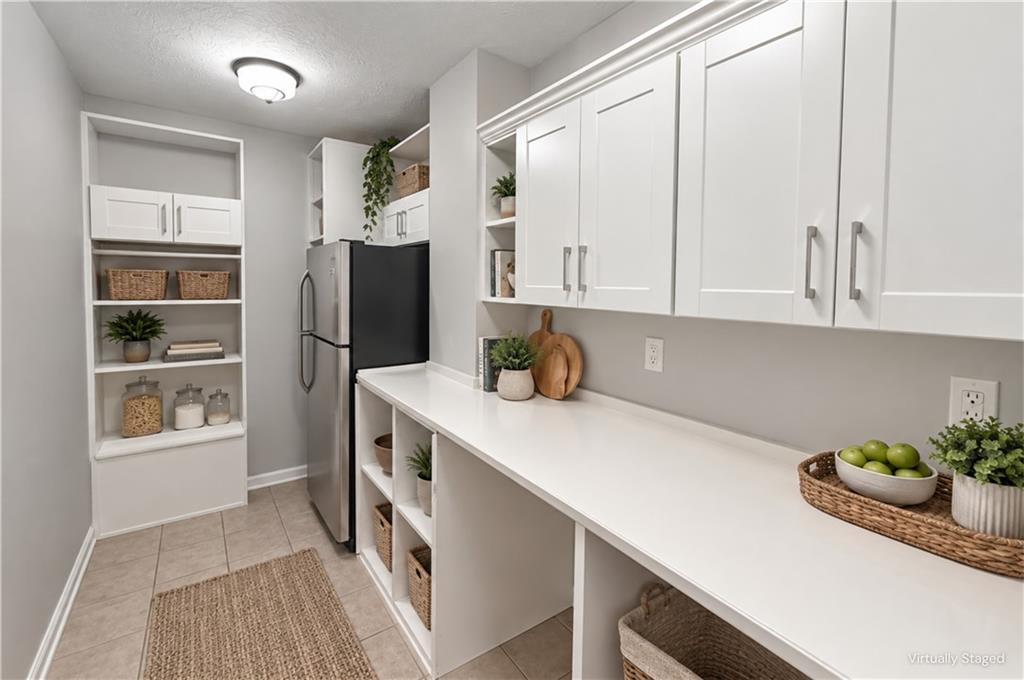 Bright staged pantry with white cabinets, a long counter, open shelving, and a stainless-steel refrigerator.
