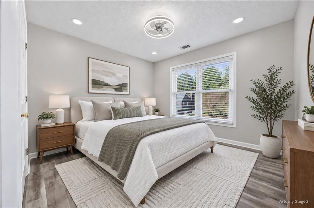 Staged bedroom with a large white bed, gray throw, wooden nightstand, and window with blinds.