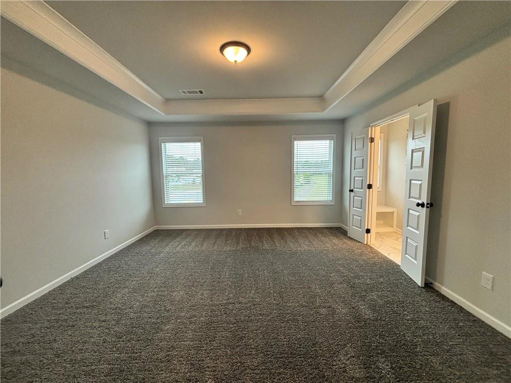 Empty carpeted room with a tray ceiling, two windows, and an open doorway to a tiled bathroom.
