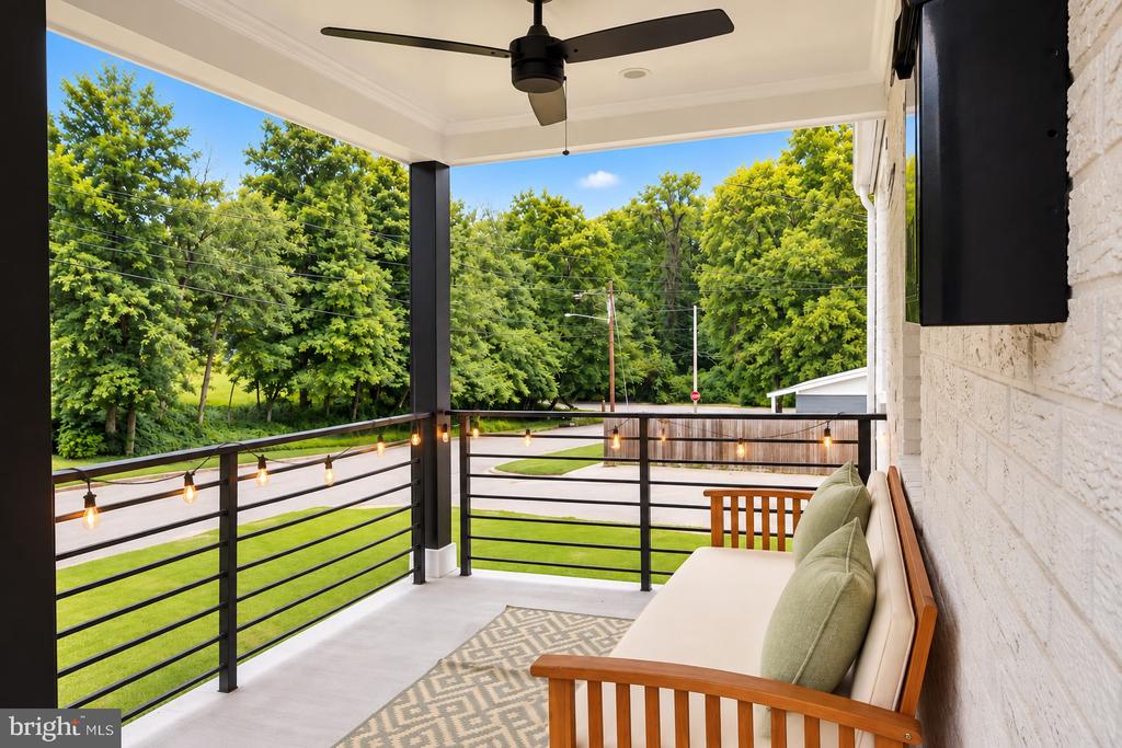Covered front porch with string lights, a ceiling fan, and a wooden bench overlooking leafy trees.